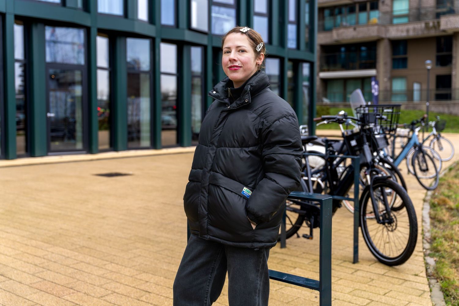 A person in black winter clothing walks through a urban setting with buildings and bicycles in the background.  - Noelle Schmeltz Fotografie