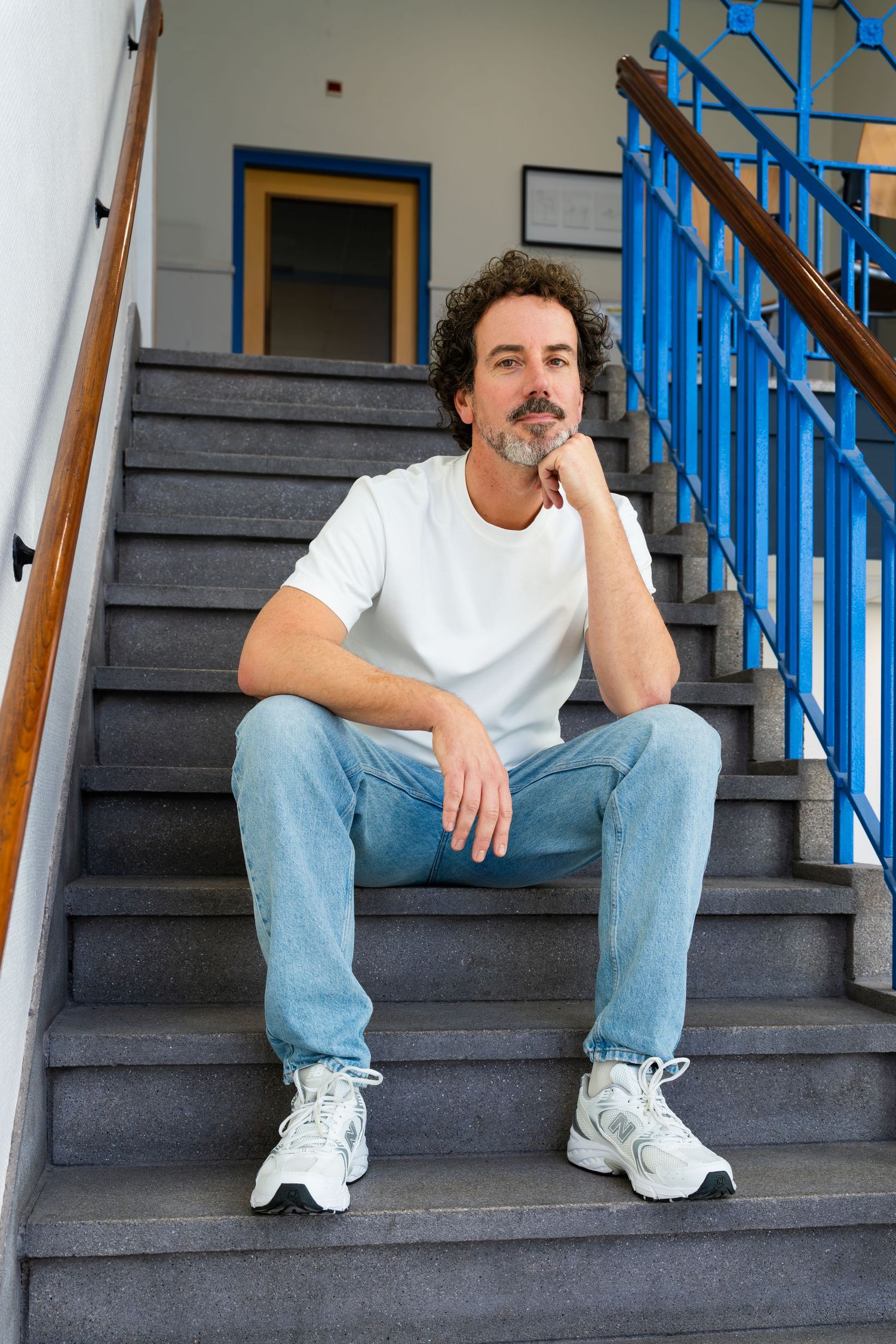 A person wearing a white t-shirt and jeans sits thoughtfully on gray concrete stairs with blue railings. - Noelle Schmeltz Fotografie