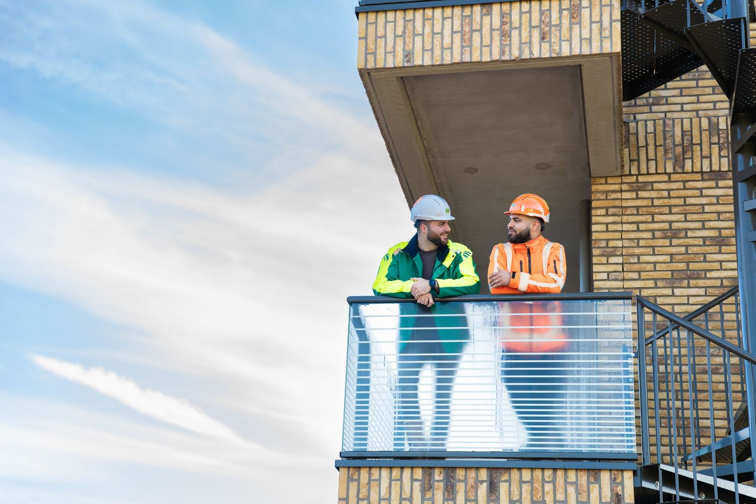 Two construction workers in high visibility safety vests stand on a balcony of a brick building against a blue sky. - Noelle Schmeltz Fotogr