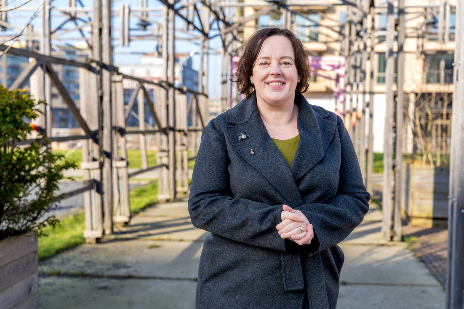 A person wearing a black coat and yellow shirt stands smiling in front of an electrical substation on a sunny day.
