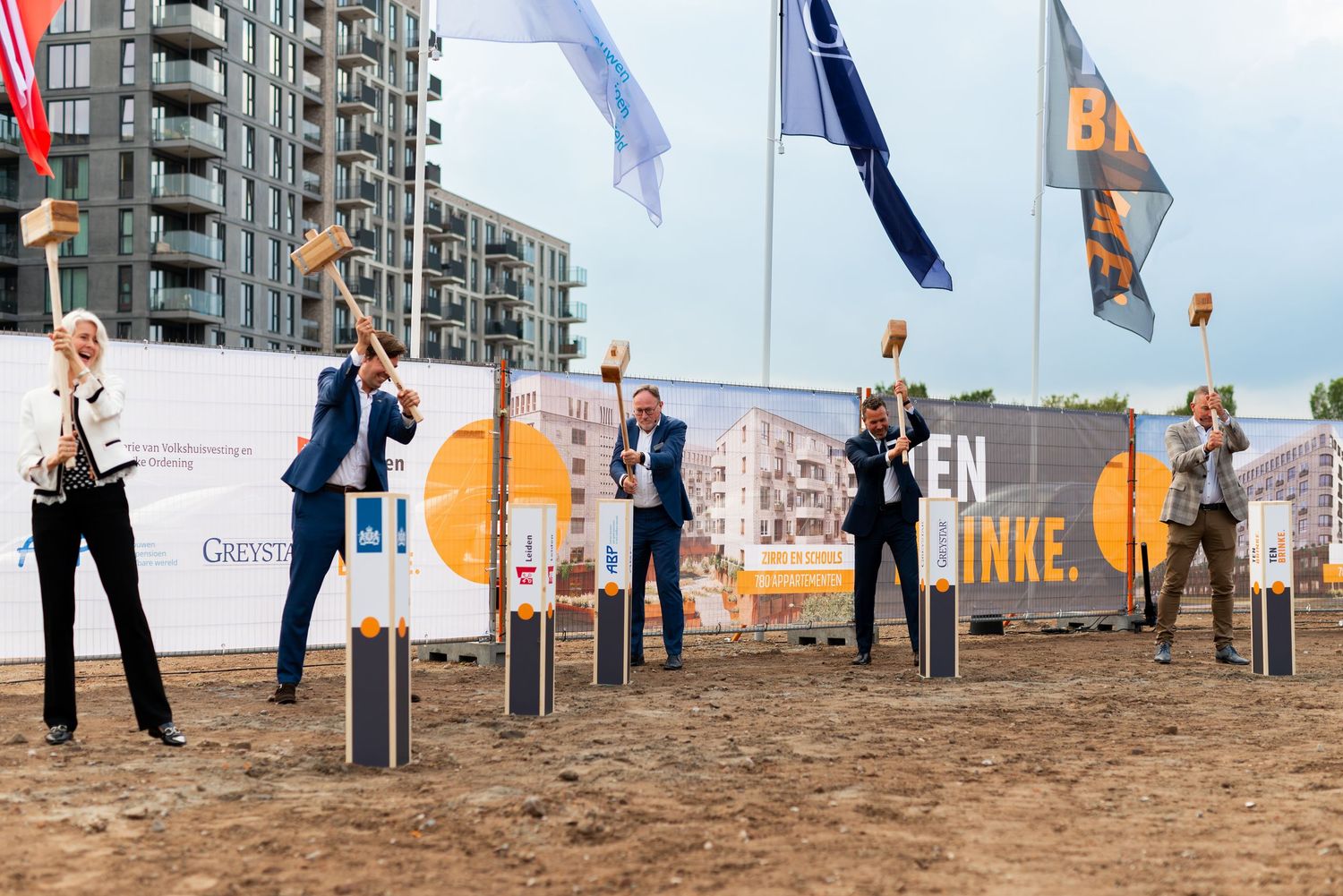 People performing ceremonial groundbreaking with shovels at construction site with flags in background. - Noelle Schmeltz Fotografie
