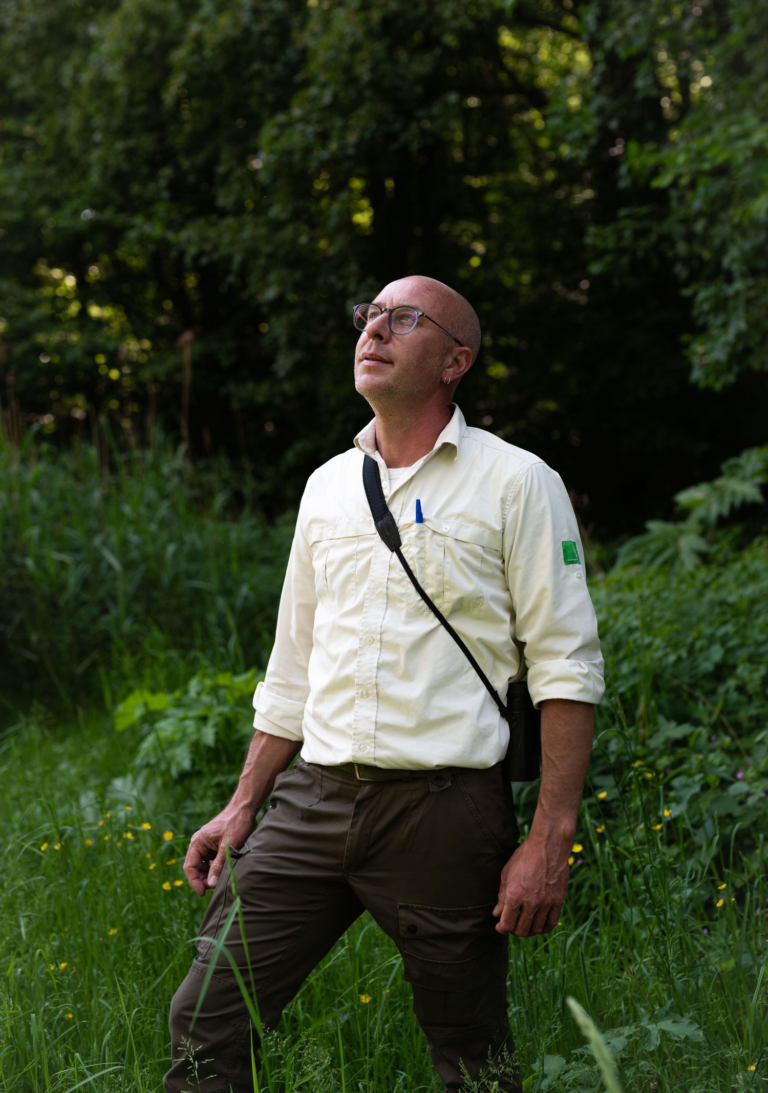 A person in a light colored shirt and dark pants stands in a lush green forest setting looking upward.
