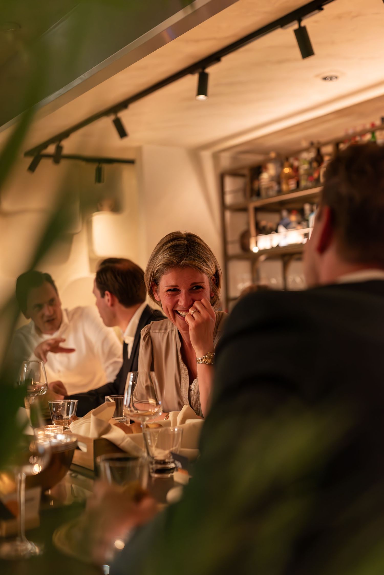 People enjoying dinner and drinks at a cozy restaurant with warm lighting and track lighting fixtures overhead. - Noelle Schmeltz Fotografie