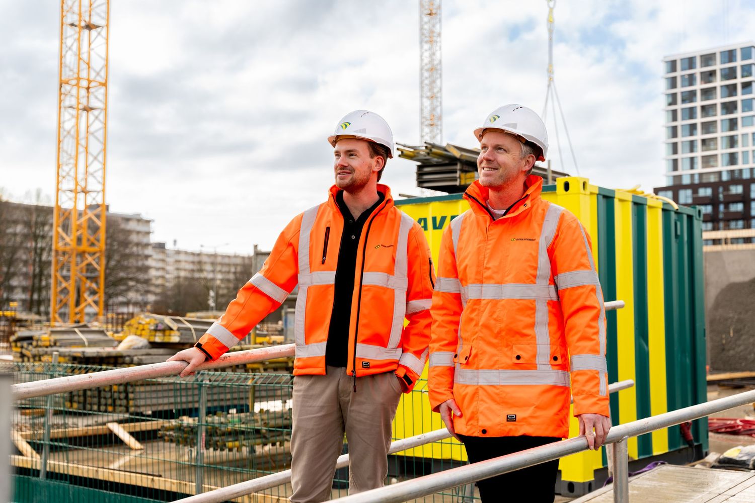 Construction workers in high visibility gear discuss plans at a building site with cranes visible in the background. - Noelle Schmeltz Fotog
