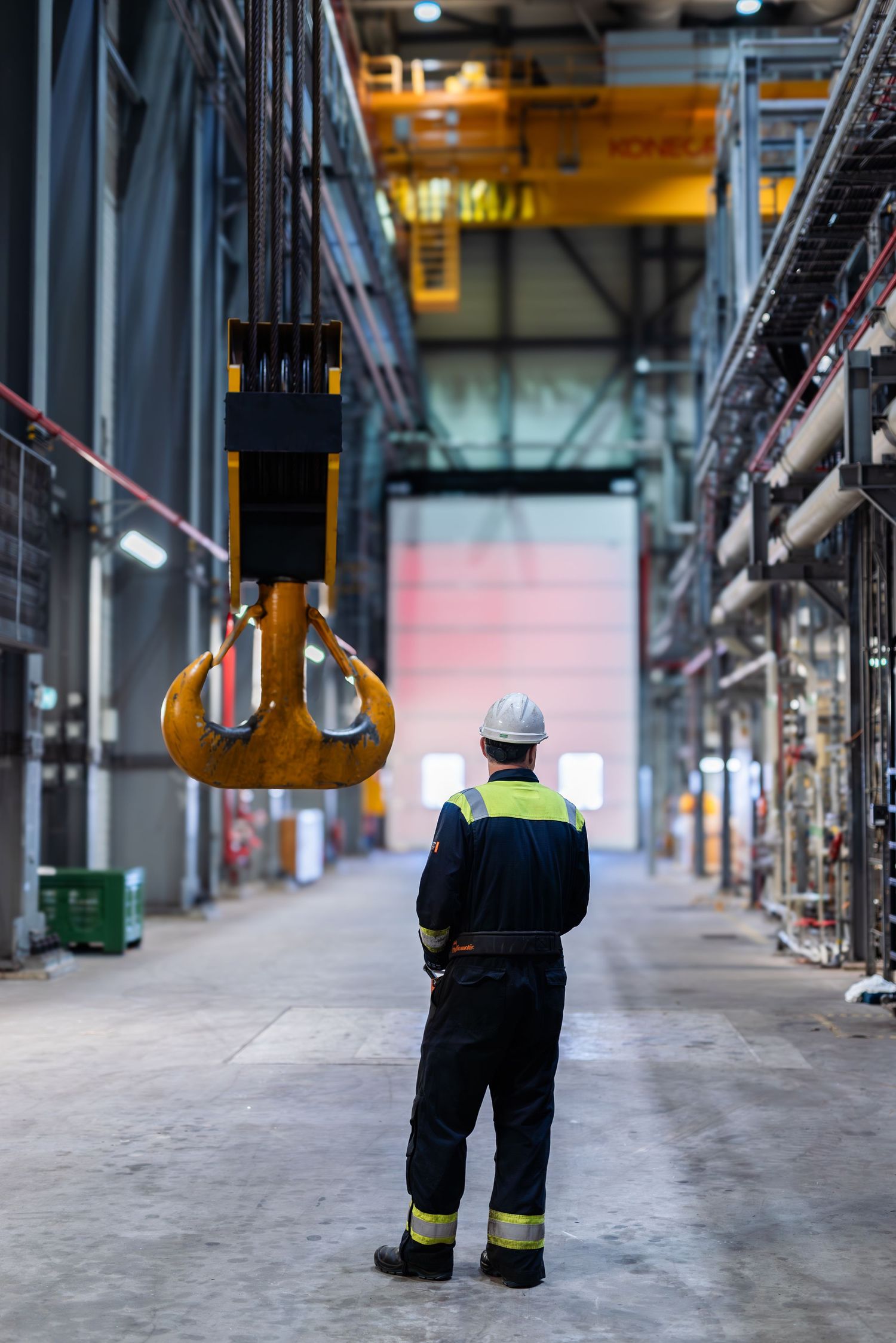 Industrial worker in safety gear and high-visibility vest walking through a large warehouse facility with metal structures.