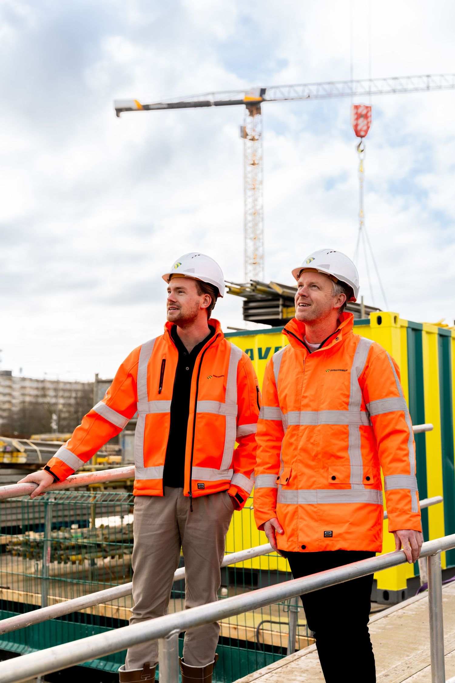 Construction workers in orange safety vests and hard hats discuss plans at a building site with cranes visible in background. - Noelle Schme