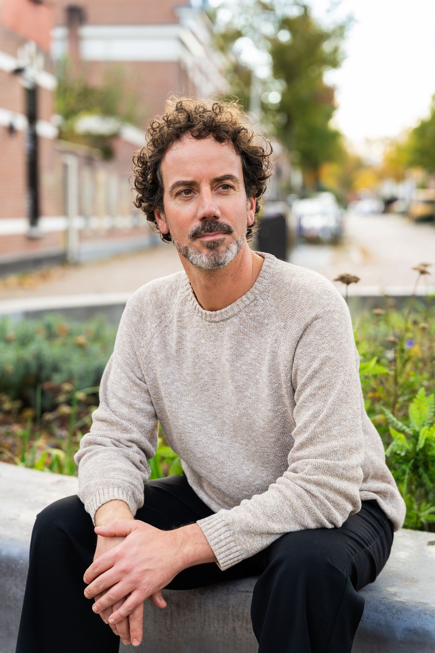 A person in a gray sweater sits casually outdoors against a blurred urban background.