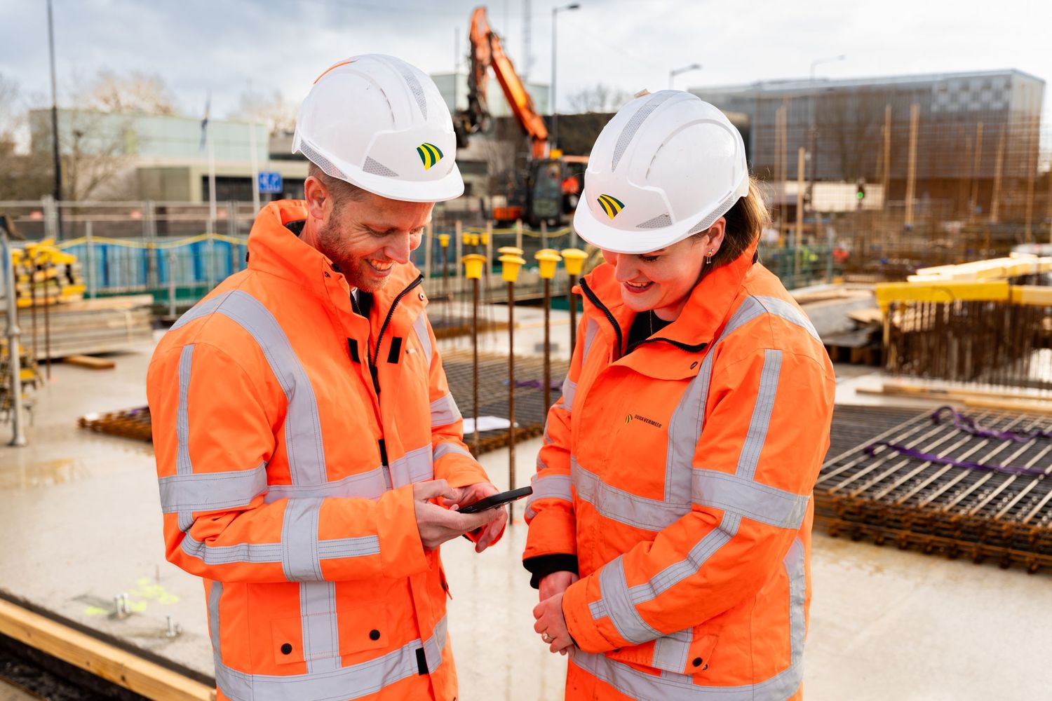 Two construction workers in orange safety gear and hard hats review information on a mobile device  - Noelle Schmeltz Fotografie