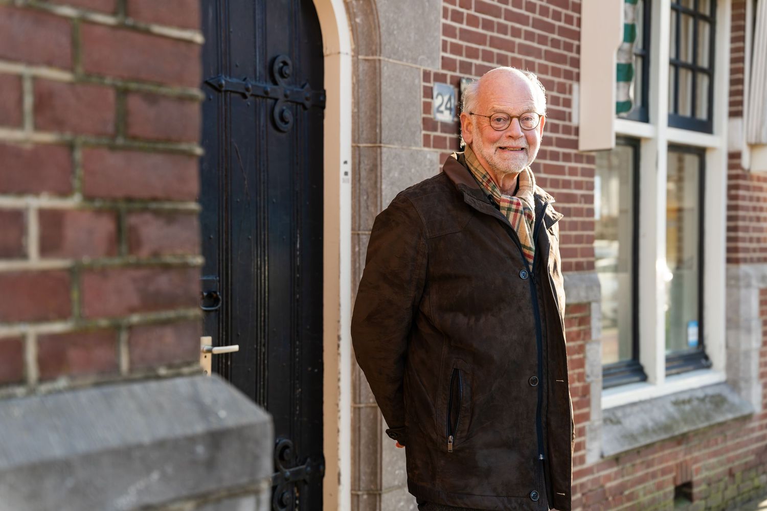 A senior man in a black coat stands in a doorway of a brick building.