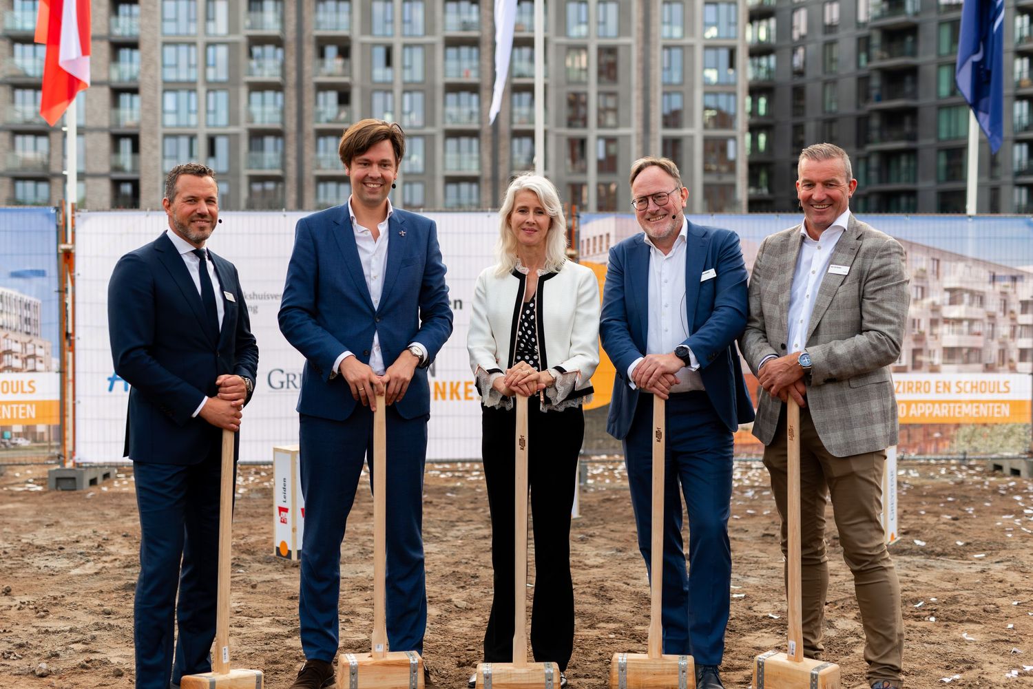 Group of people with shovels at groundbreaking ceremony in front of apartment buildings. - Noelle Schmeltz Fotografie