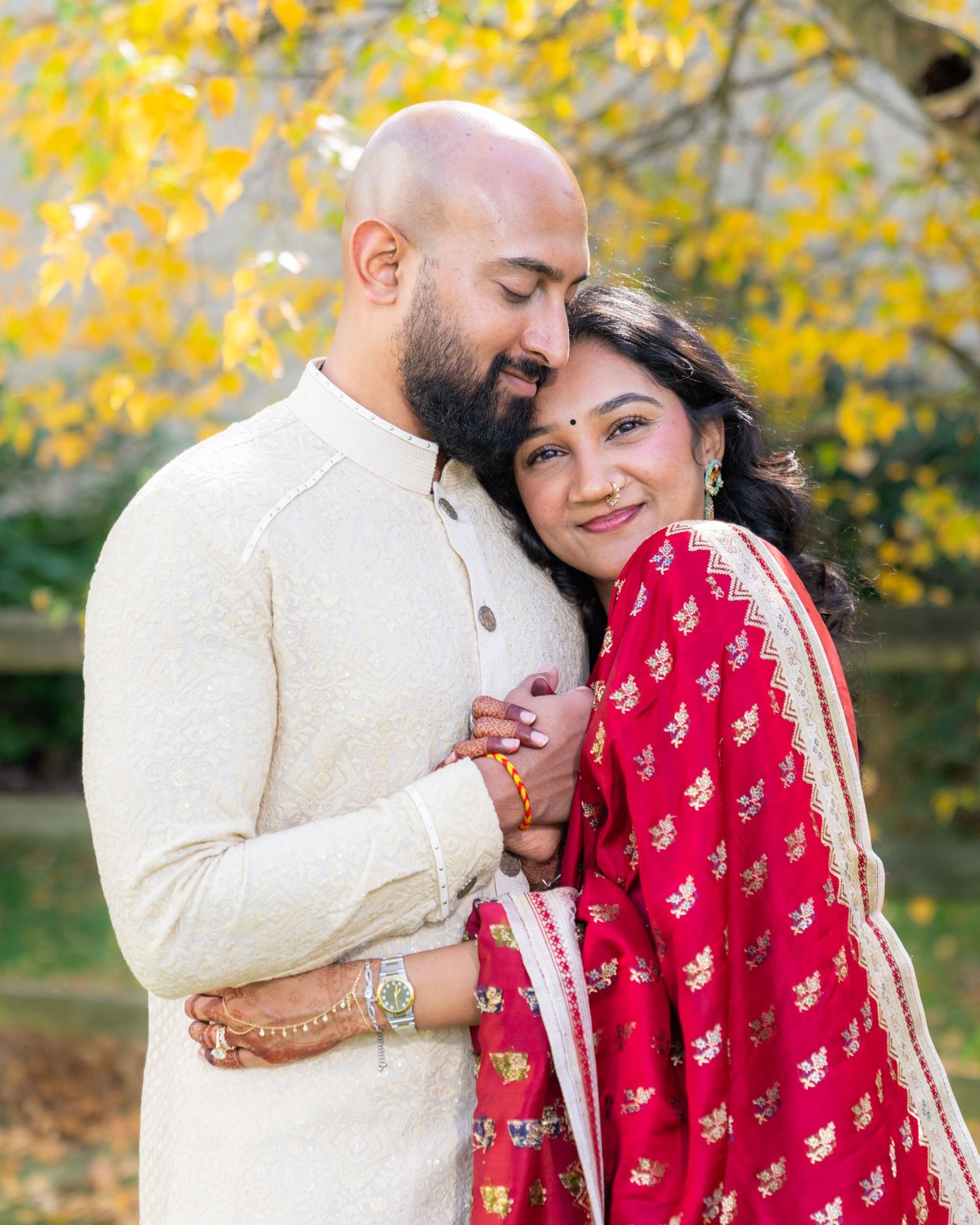 A couple in traditional Indian wedding attire share an intimate moment against a backdrop of yellow autumn foliage.
