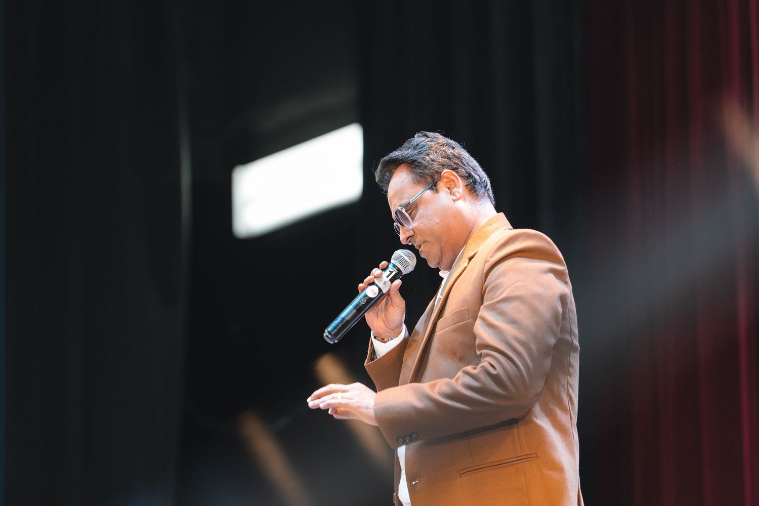 A singer performs on stage wearing a dark suit while holding a microphone against a dark background.