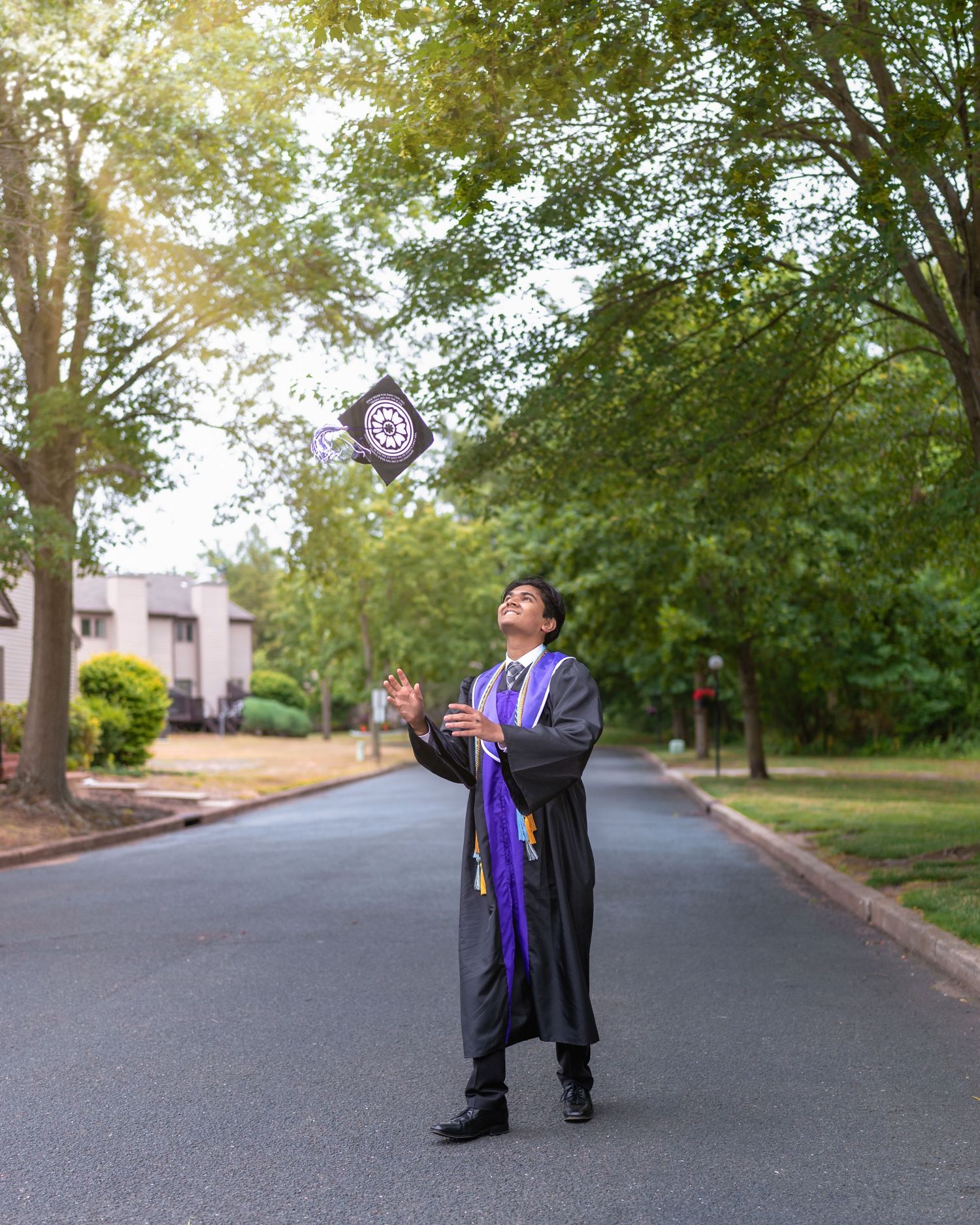 Graduate tossing cap into air on campus road surrounded by trees.