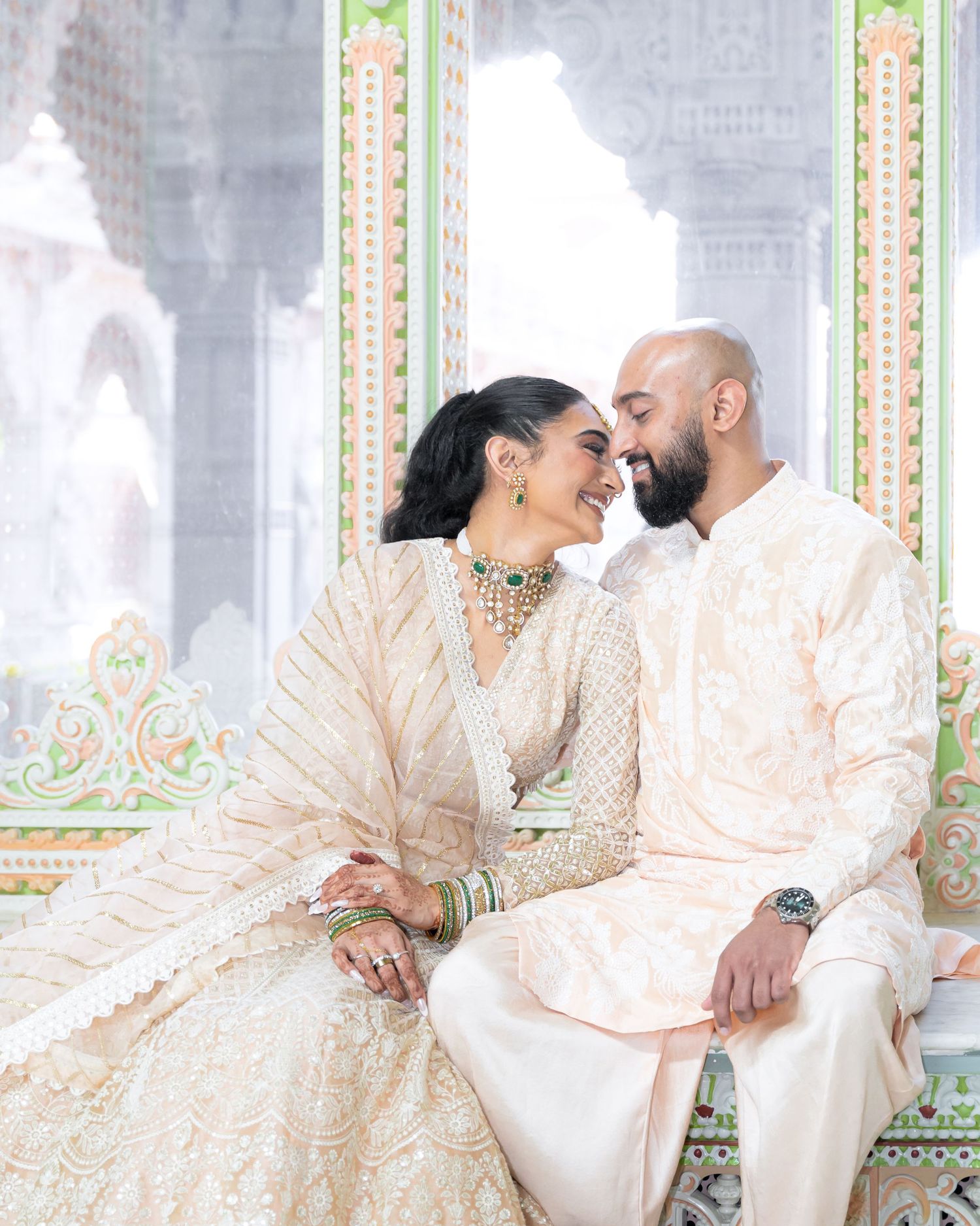 A couple in elegant cream-colored traditional attire share an intimate moment on an ornate bench.
