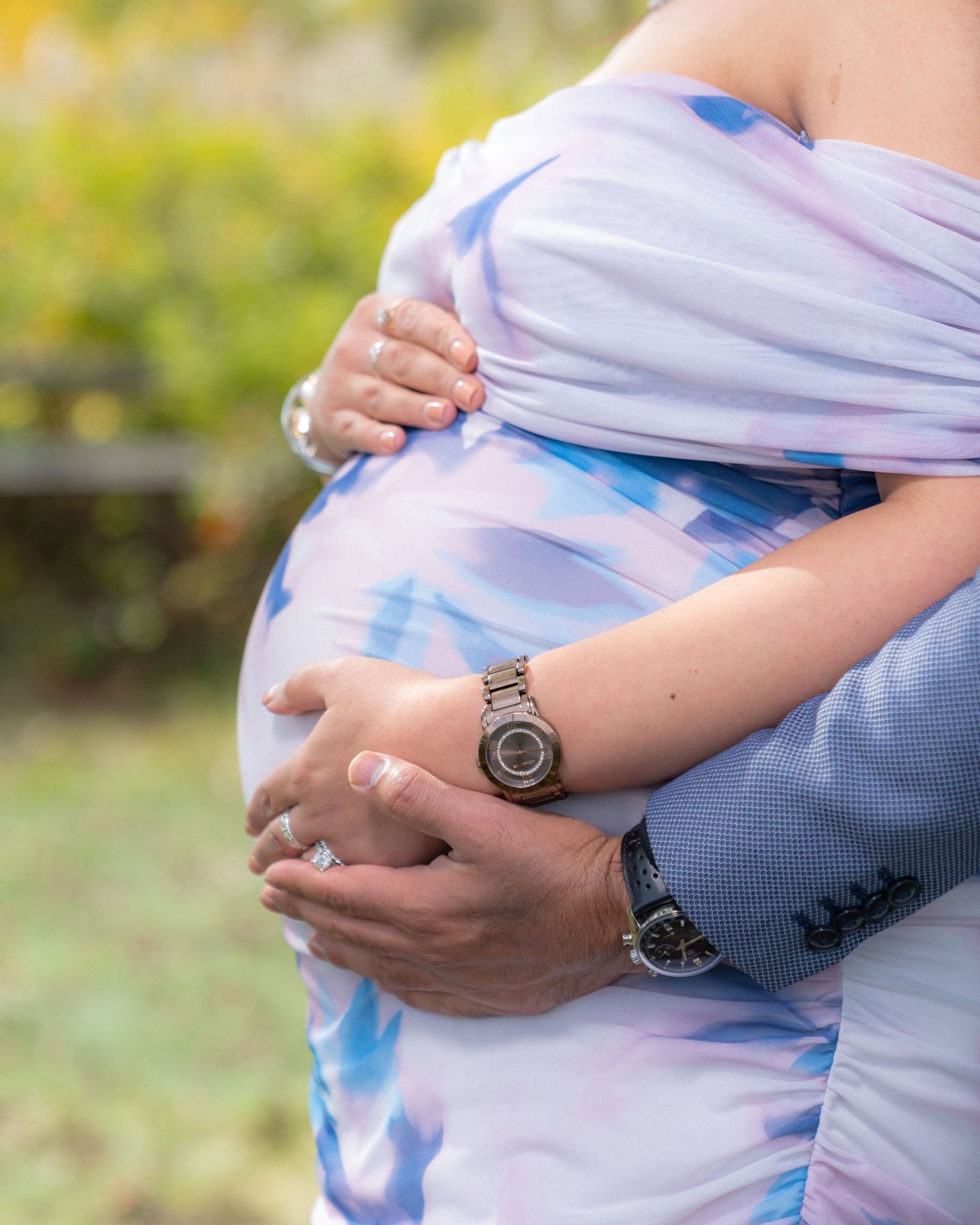 Hands gently cradle a pregnant belly wearing a light blue and white tie-dye maternity dress outdoors.