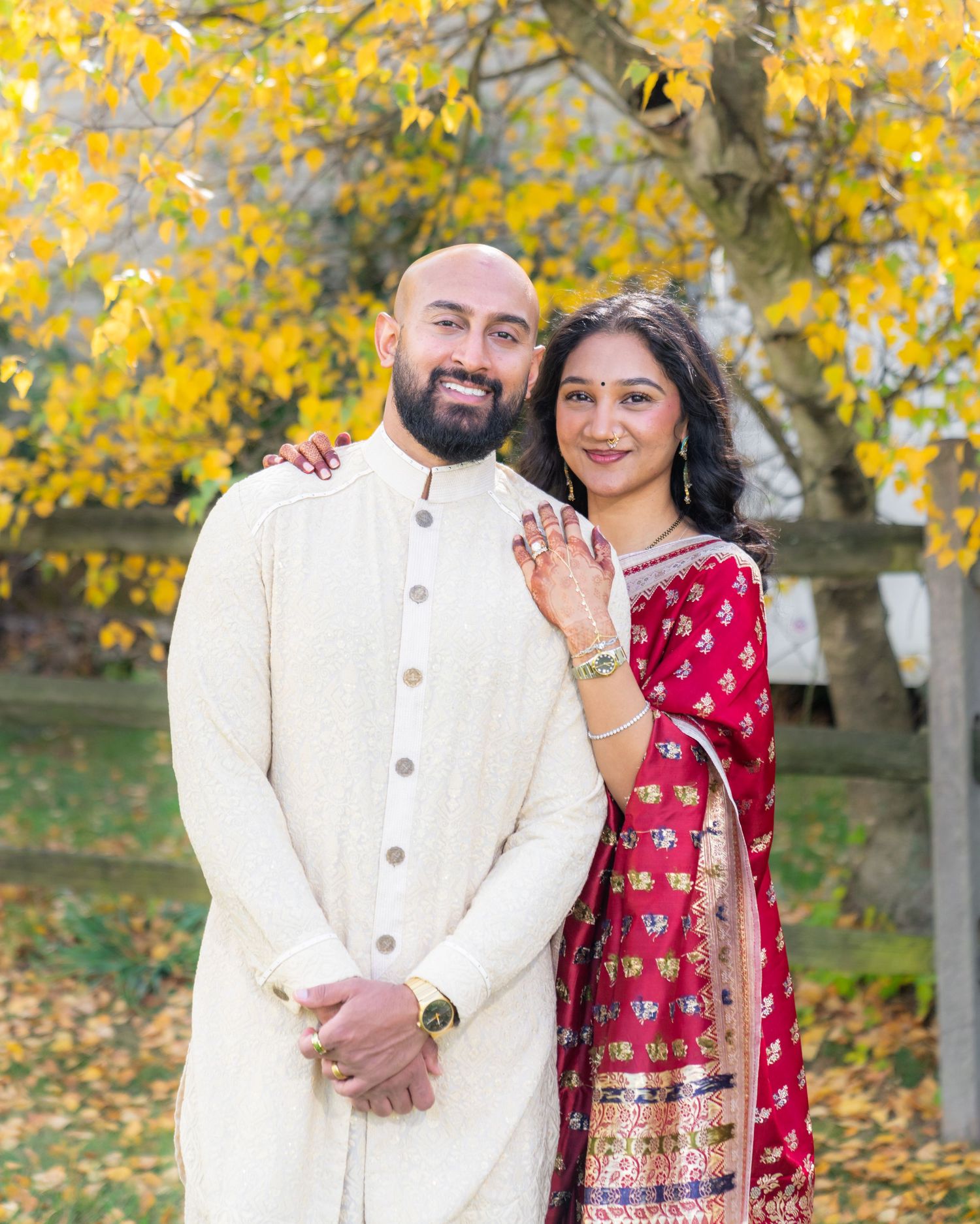 A couple in traditional South Asian attire poses outdoors with vibrant yellow autumn foliage in the background.