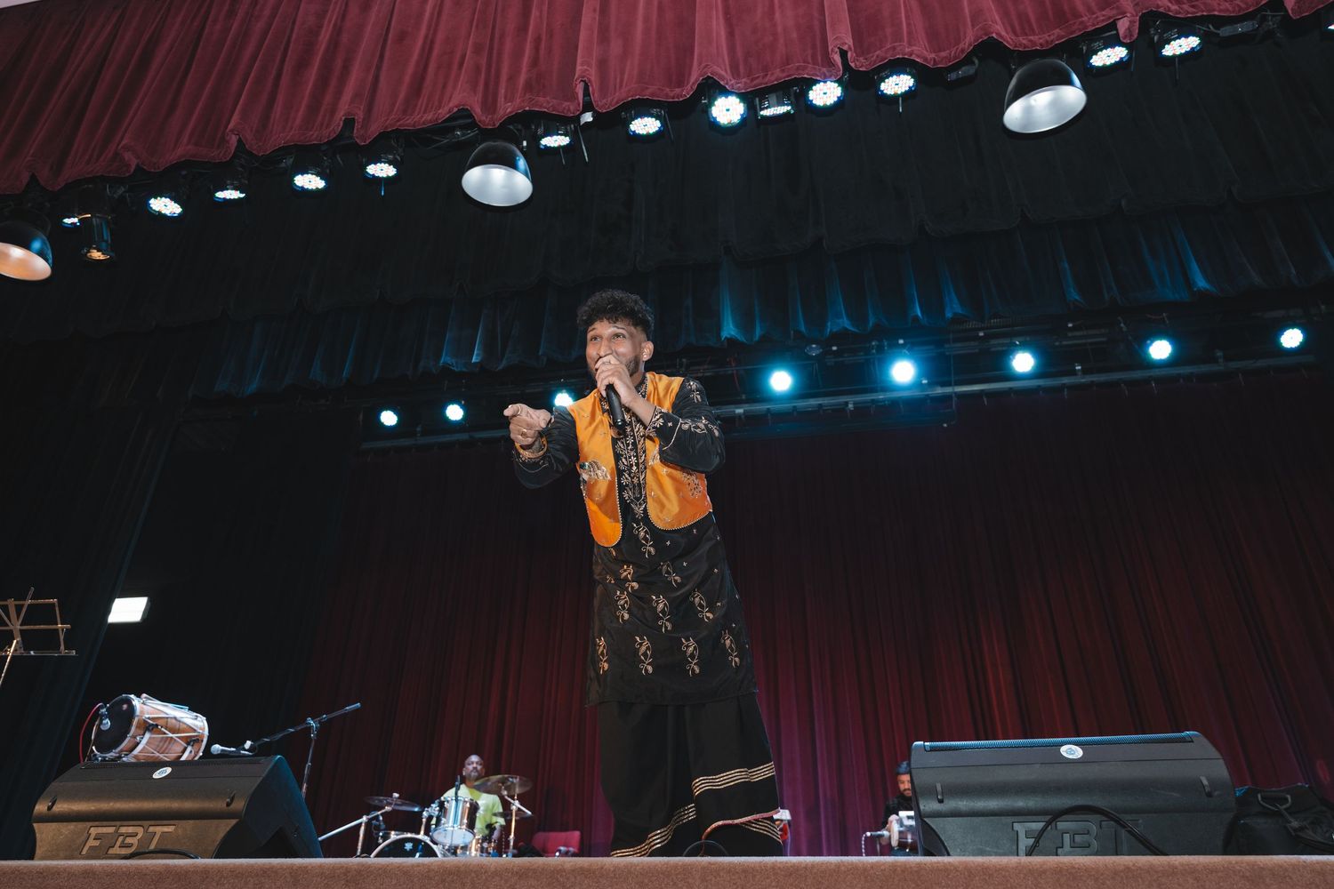 Concert performer in bright yellow vest energetically singing on dimly lit stage with red curtains.