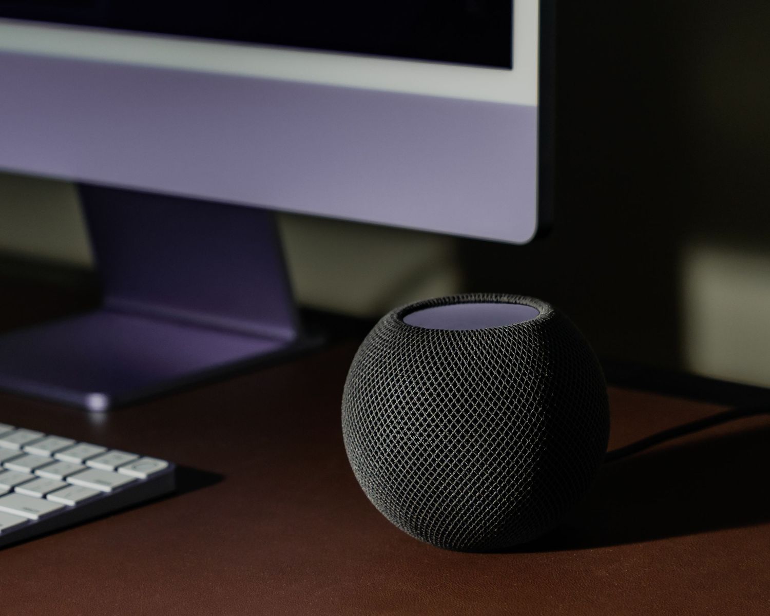 HomePod Mini speaker sitting on desk next to laptop keyboard in dark ambient lighting.