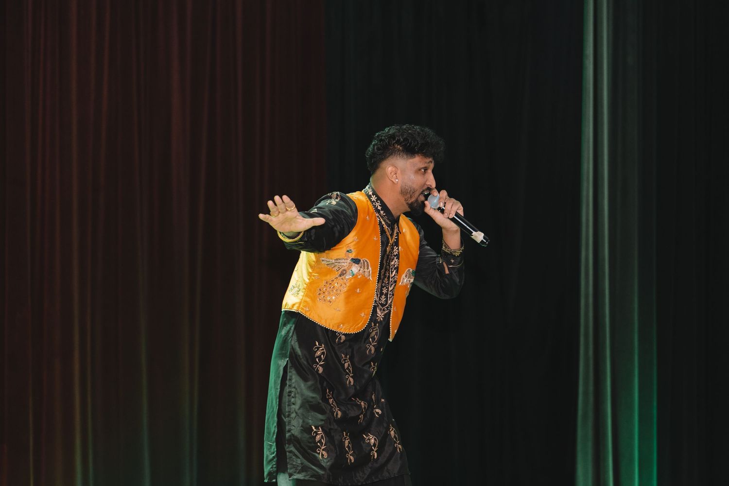 Performer in orange jacket singing on stage with dramatic red curtain backdrop and theatrical lighting.