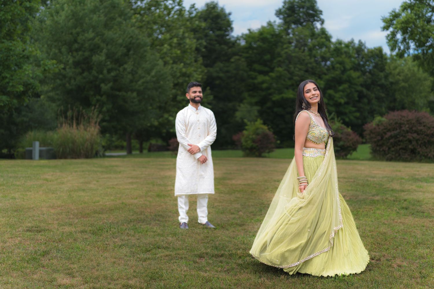 Couple in traditional Indian attire posing outdoors, woman in yellow lehenga and man in white kurta, lush green park background.
