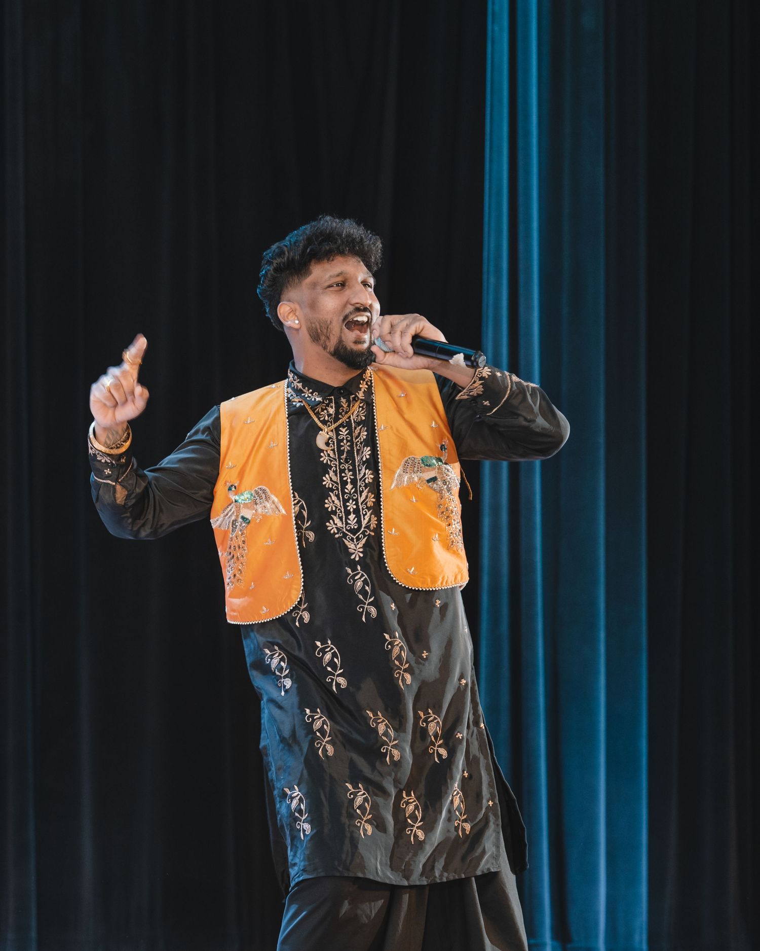 Performer in black and gold vest energetically singing on stage with dramatic blue lighting and theatrical gestures.