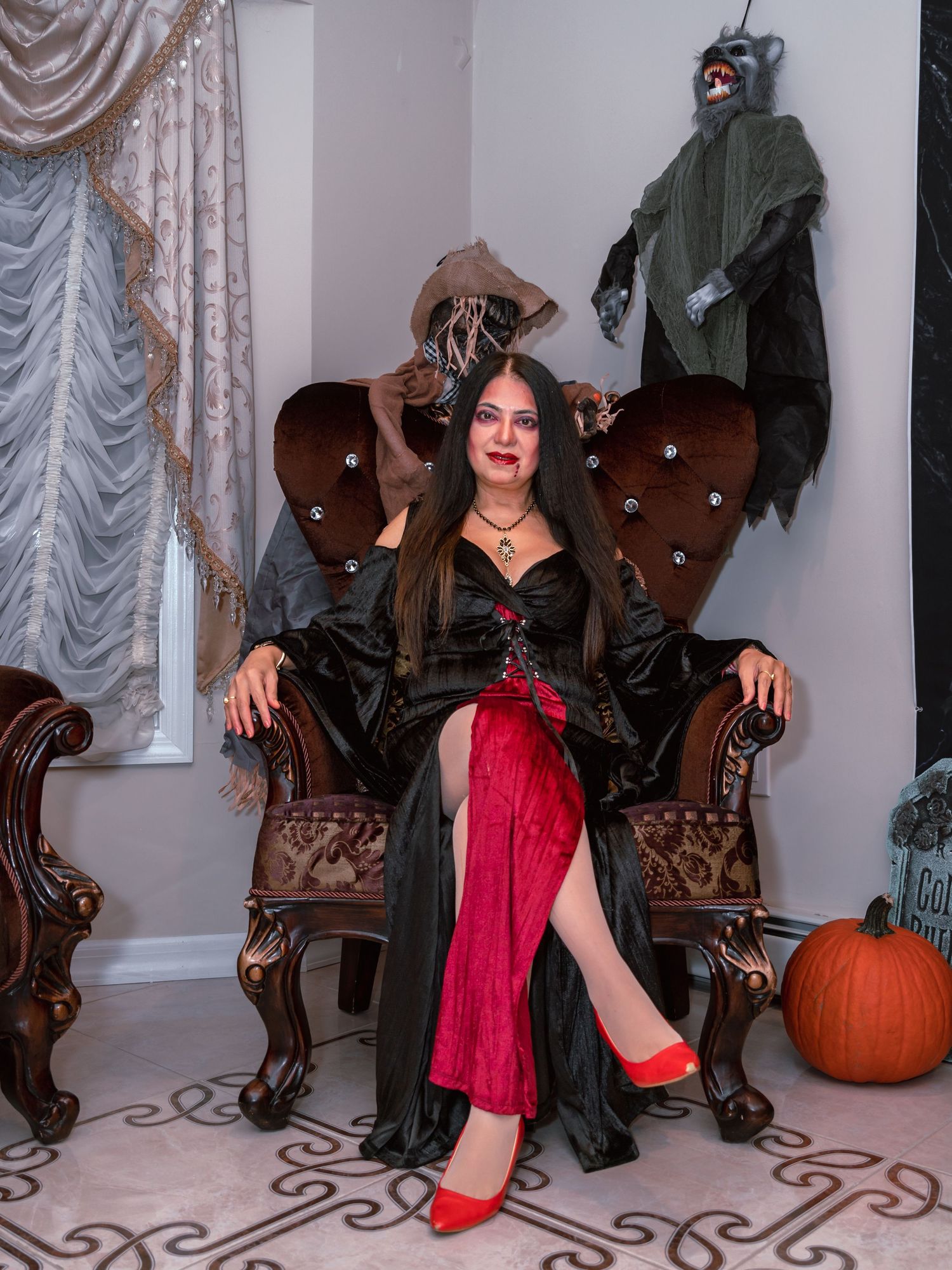 A gothic-themed photoshoot featuring a model in black and red attire posing on an ornate chair with Halloween decor.