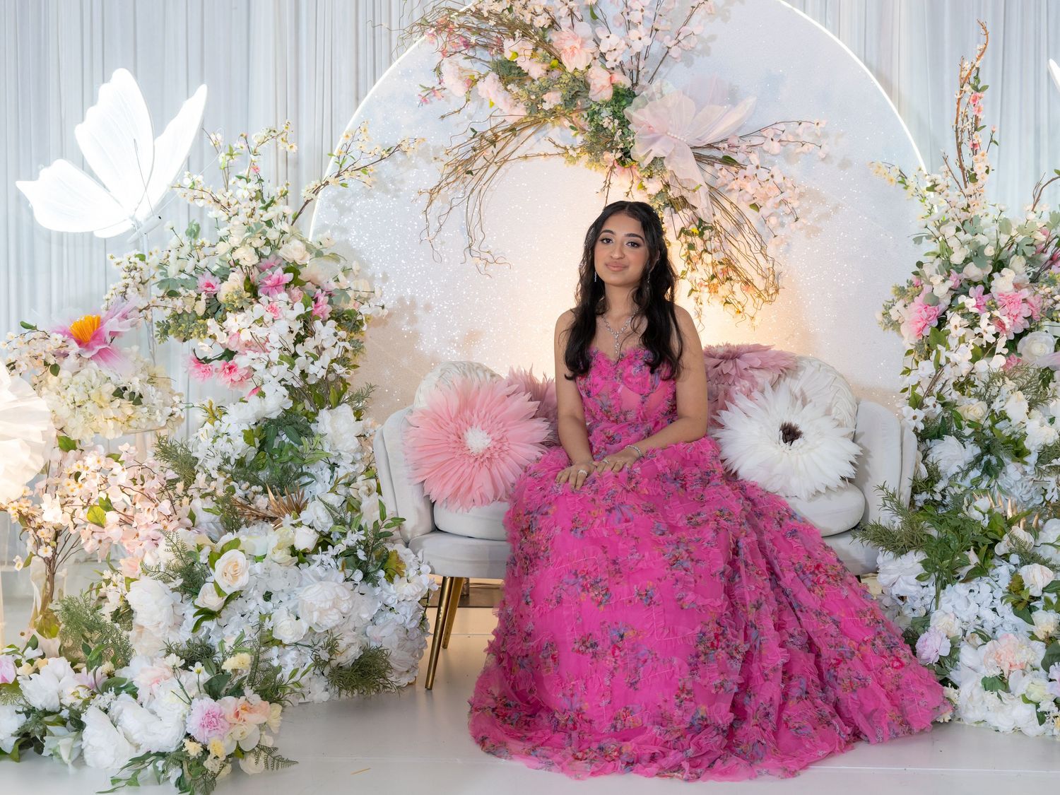 A person in a vibrant hot pink ball gown poses elegantly among white floral arrangements and spring blossoms.
