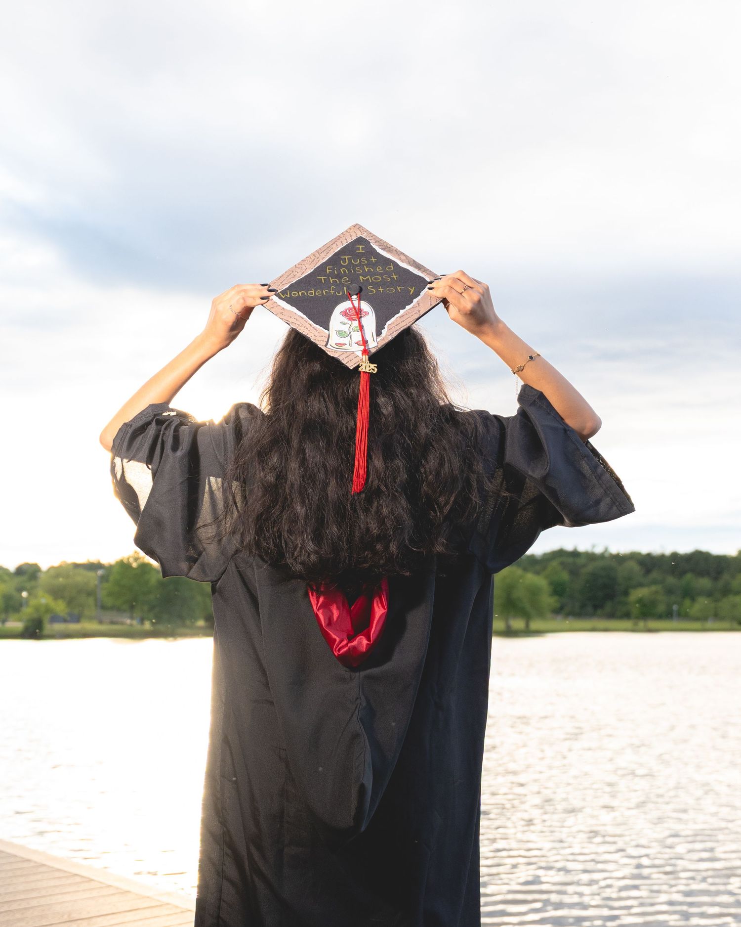 Graduate in black gown holding red tassel against lake backdrop.