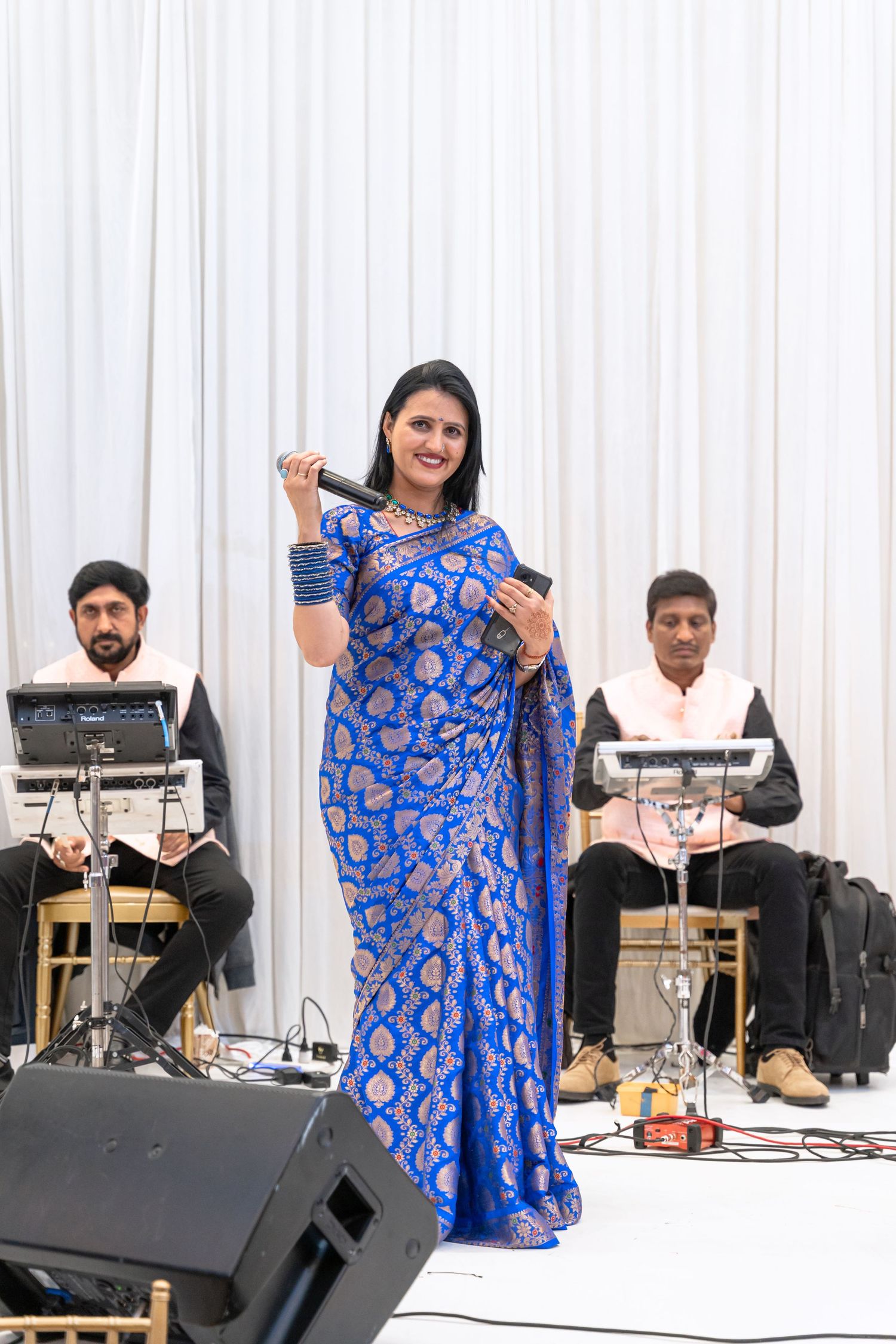 Musicians perform on stage with a singer in a blue patterned dress accompanied by tabla and keyboard players.