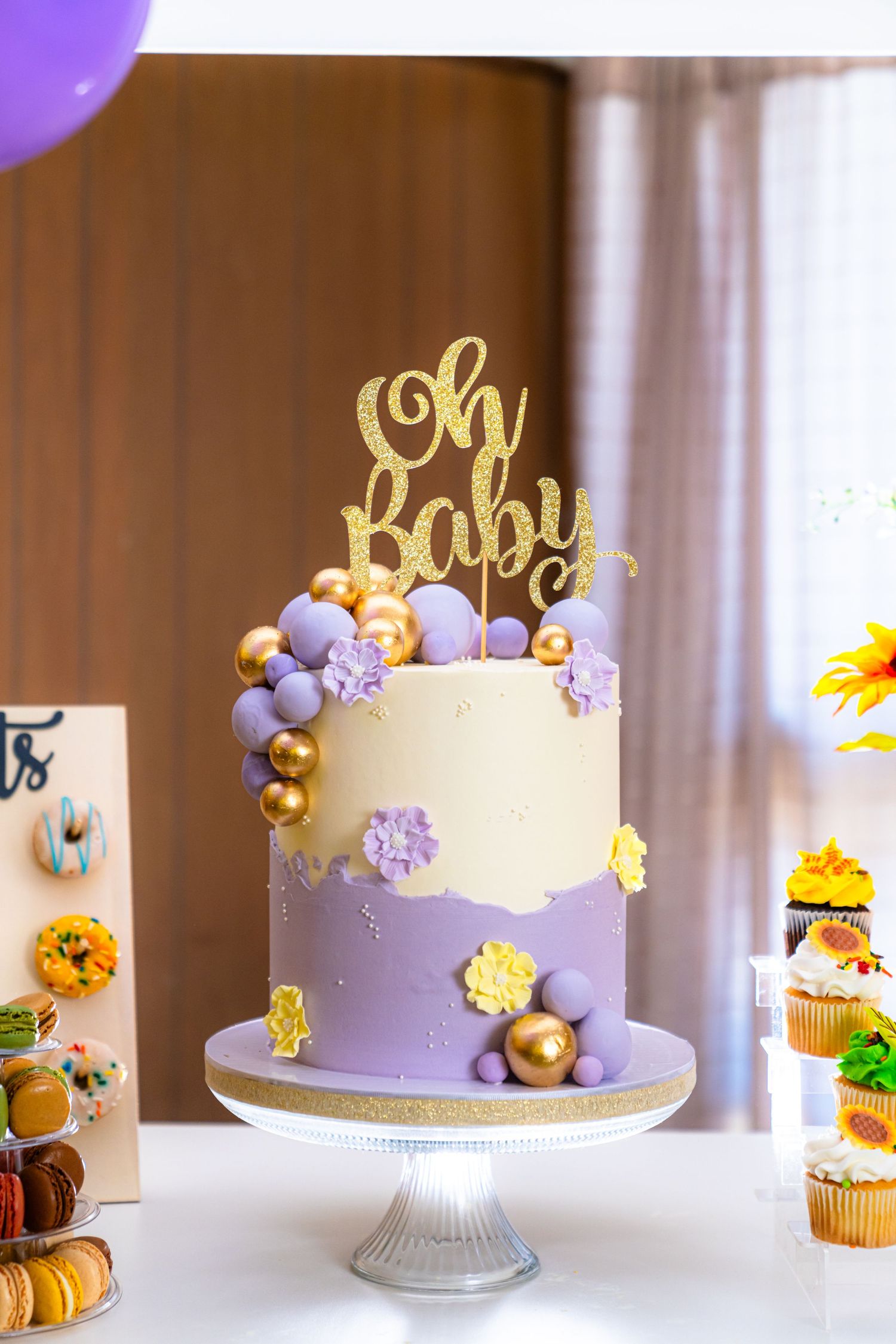 Two-tier baby shower cake with gold topper and purple flowers on white cake stand.