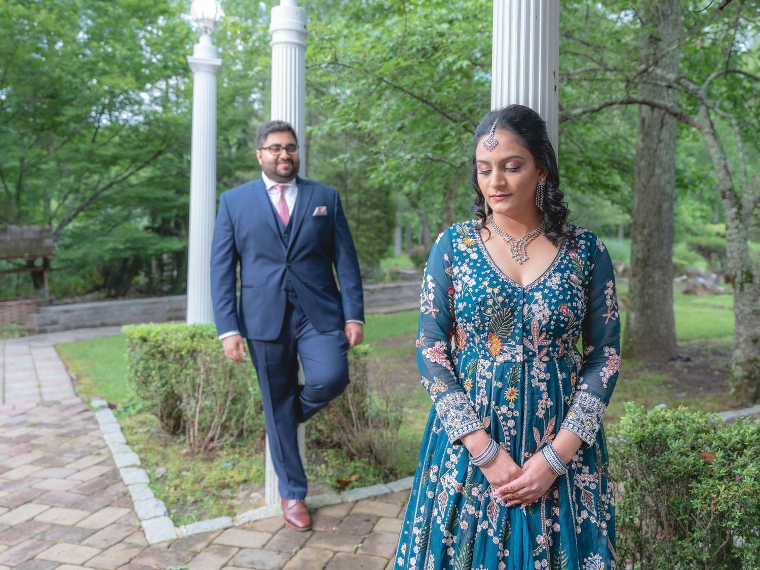 Couple walking through a garden path lined with classical columns and trees.