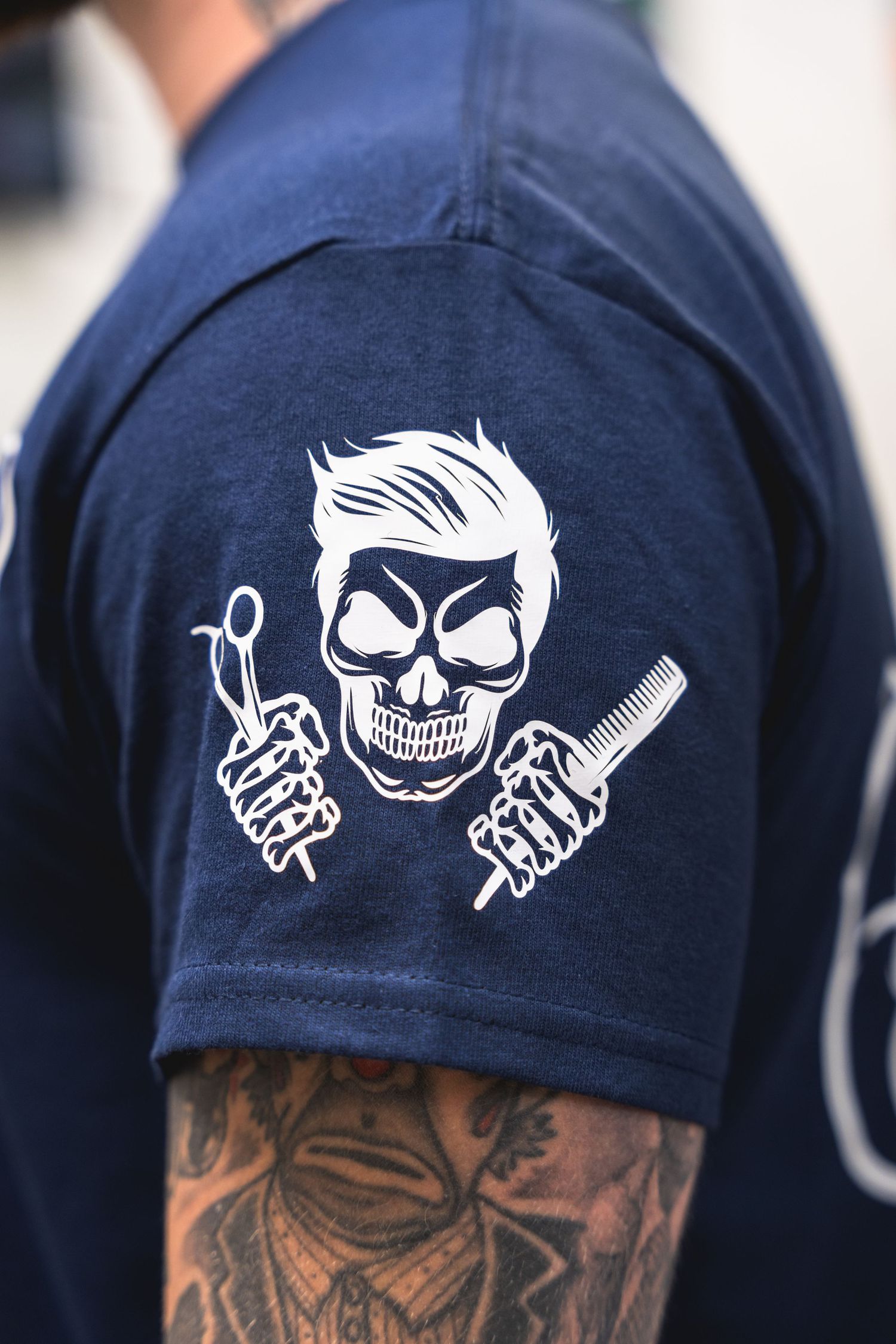 Navy blue t-shirt sleeve showing a cartoon skull barber design with hair styling tools.