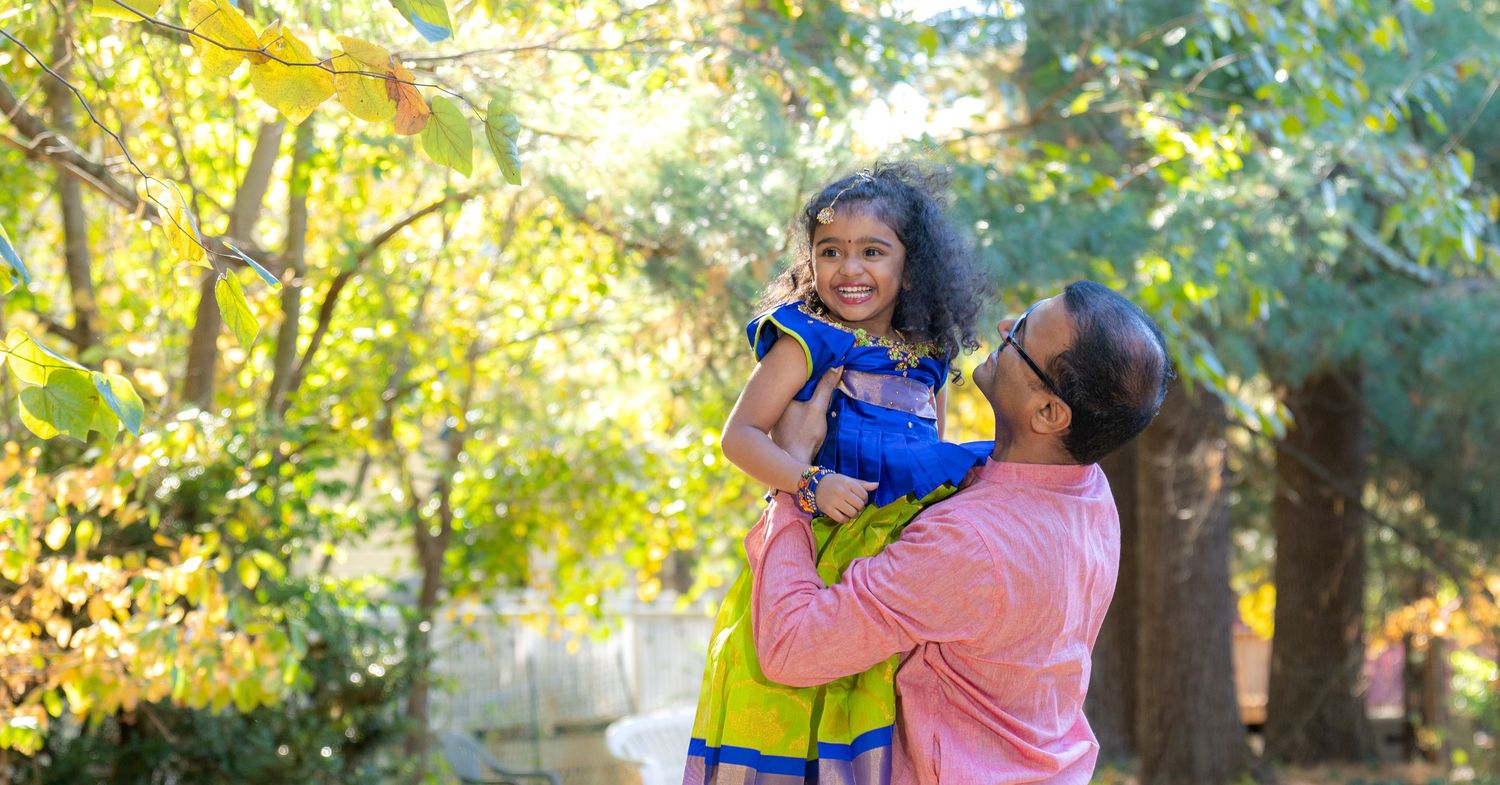 A parent and child share a joyful moment together outdoors in a sunlit park setting with autumn trees in the background.