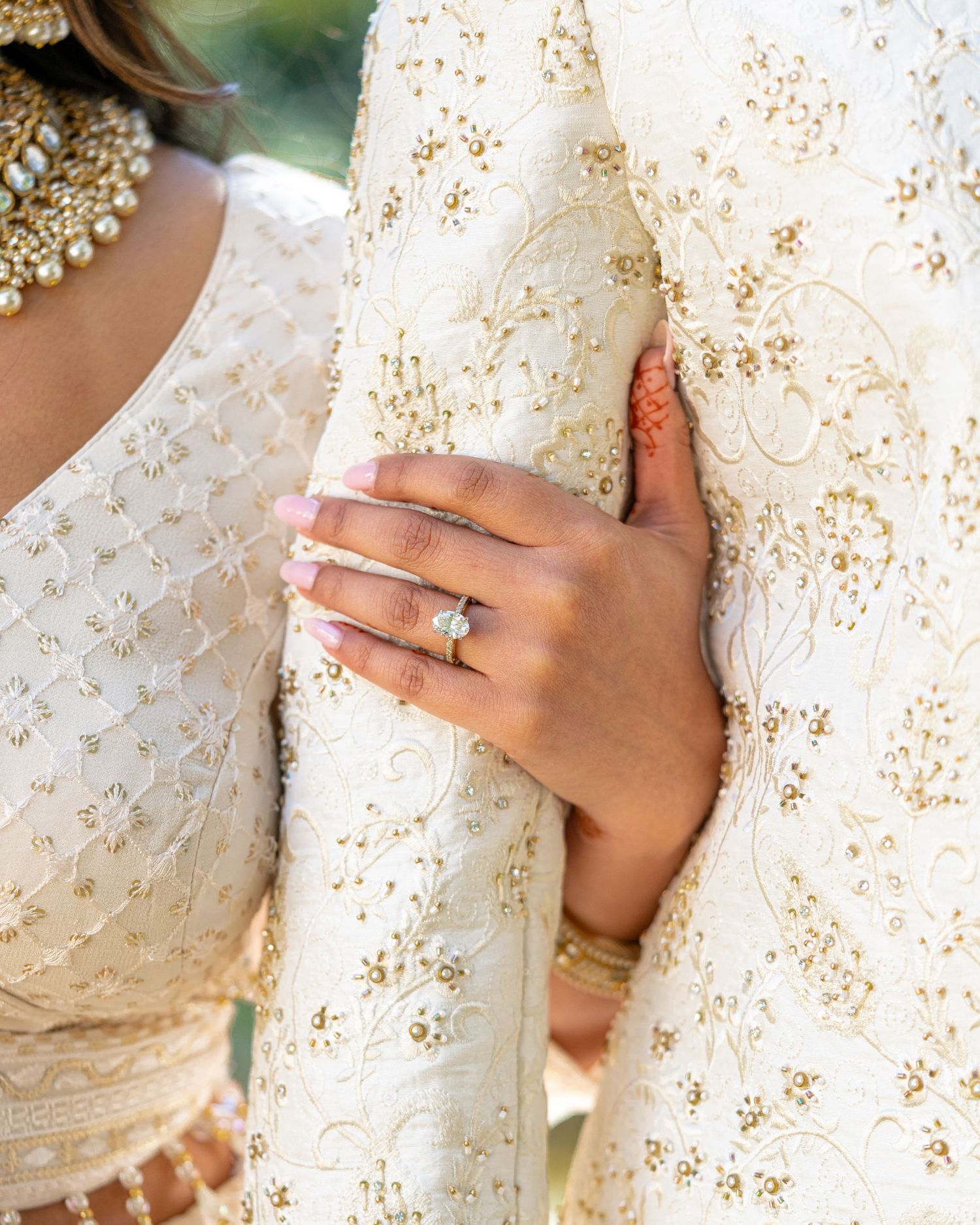 A close-up of an ornate engagement ring on a hand against white and gold embroidered fabric.