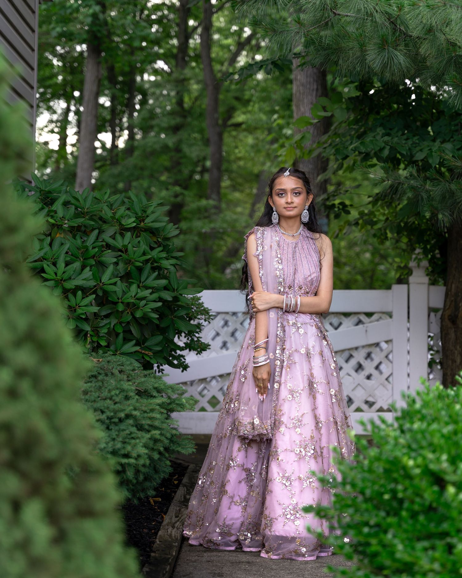 Woman wearing pink floral lehenga poses in garden with white lattice backdrop.