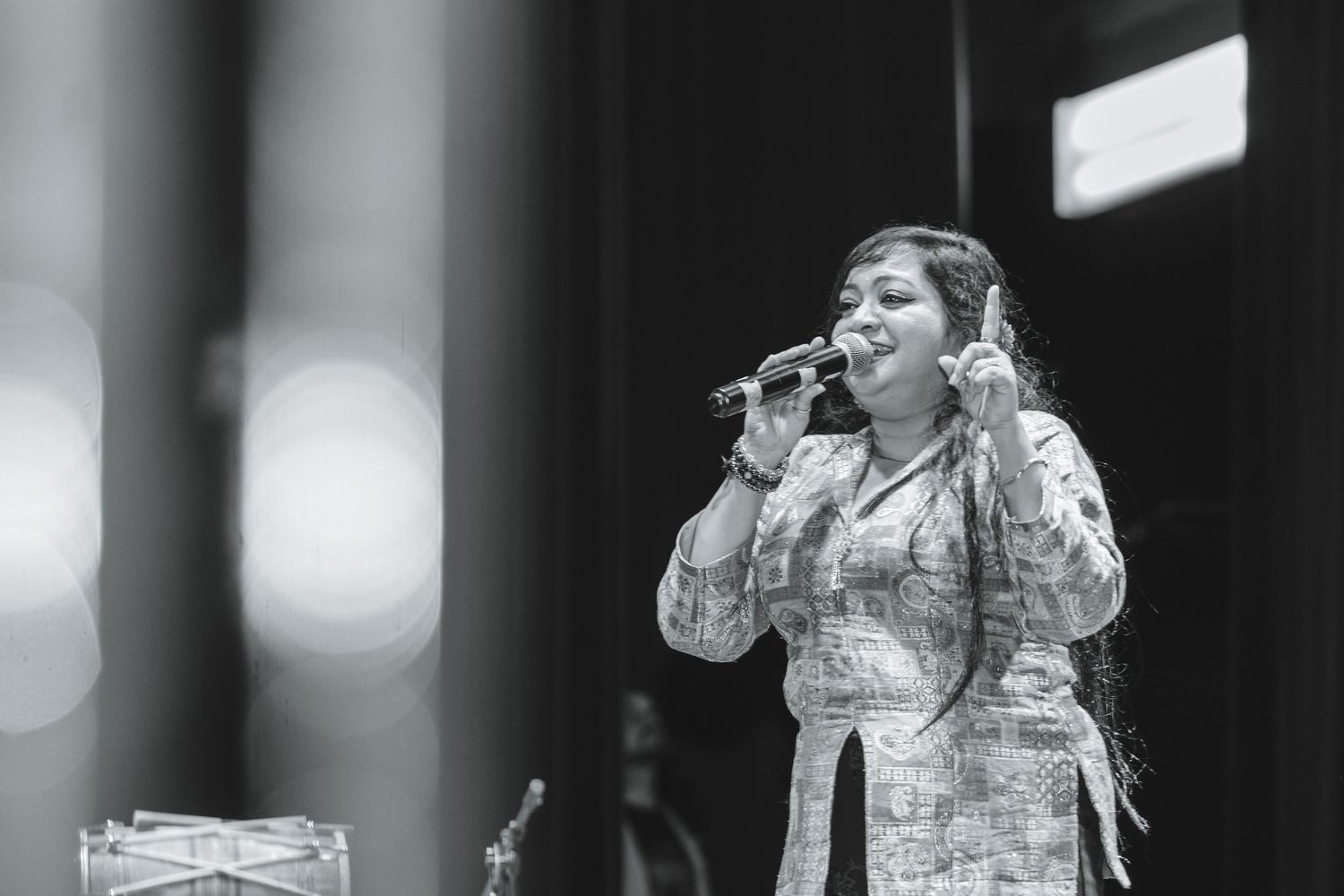 A performer in a glittering white outfit sings passionately on stage with dramatic lighting effects in the background.