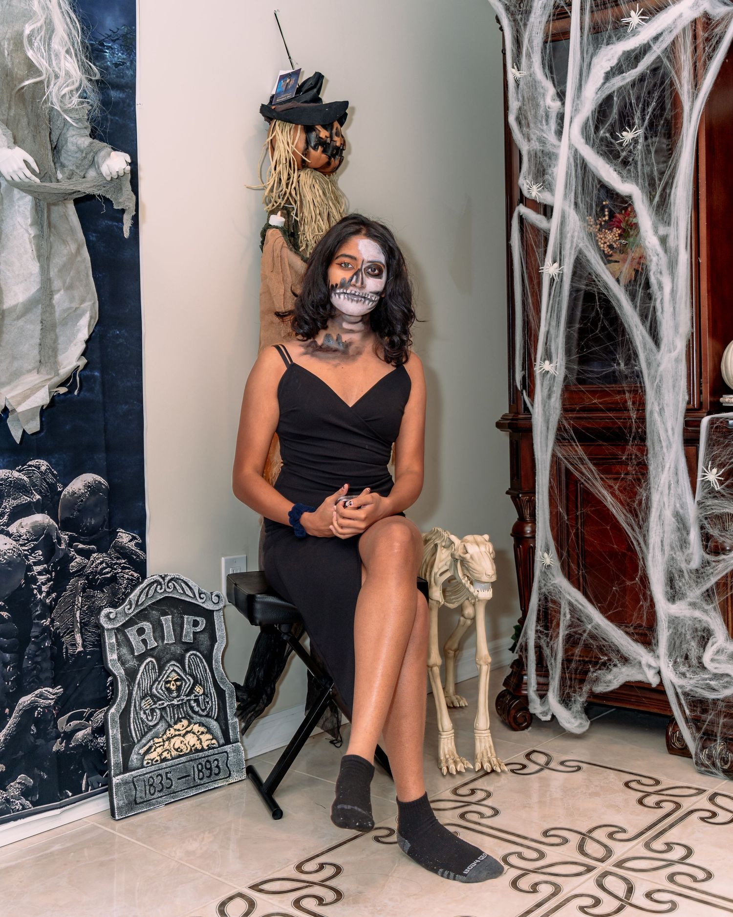 A person in a black dress and Halloween-themed accessories poses next to spooky white cobweb decorations on a wall.