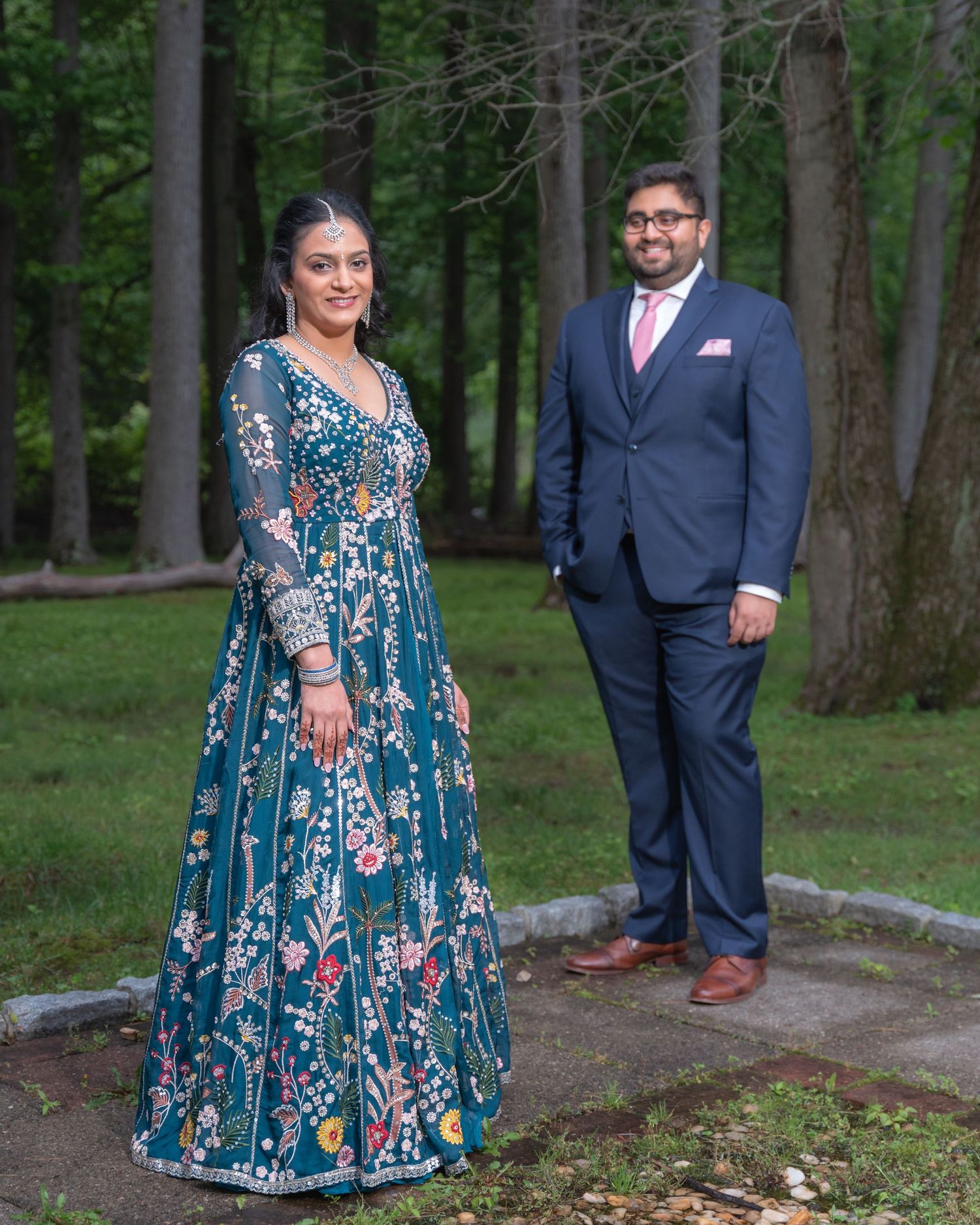 A couple poses in an outdoor garden setting, with one wearing a floral blue dress and the other in a navy suit.