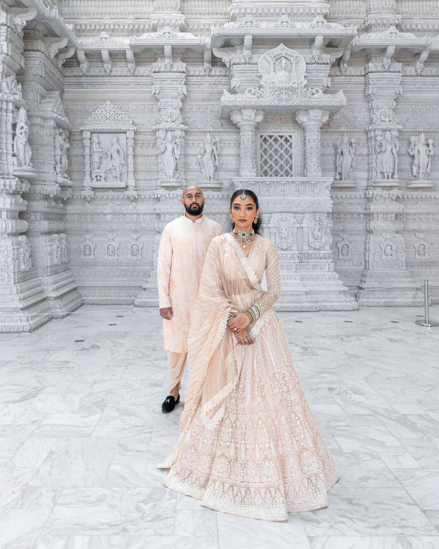 A couple in cream-colored traditional outfits walking near an intricately carved white temple wall.