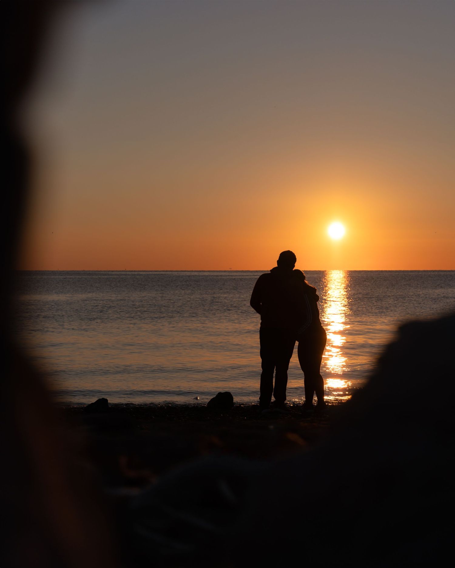 Couple's silhouette standing together watching sunset over peaceful ocean waters.