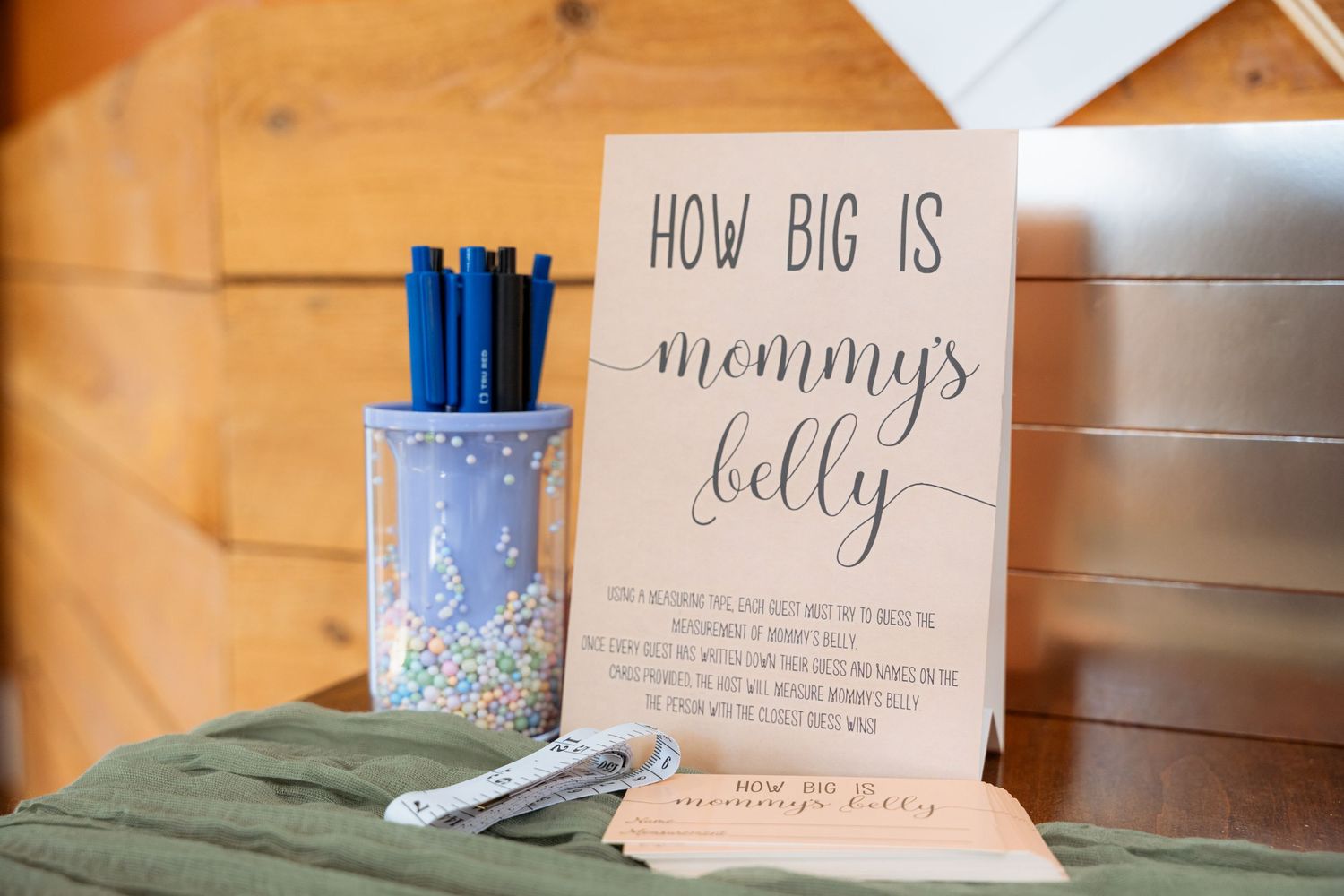 A wooden table displays a 'How big is mommy's belly' sign next to a floral pen holder with blue pens.