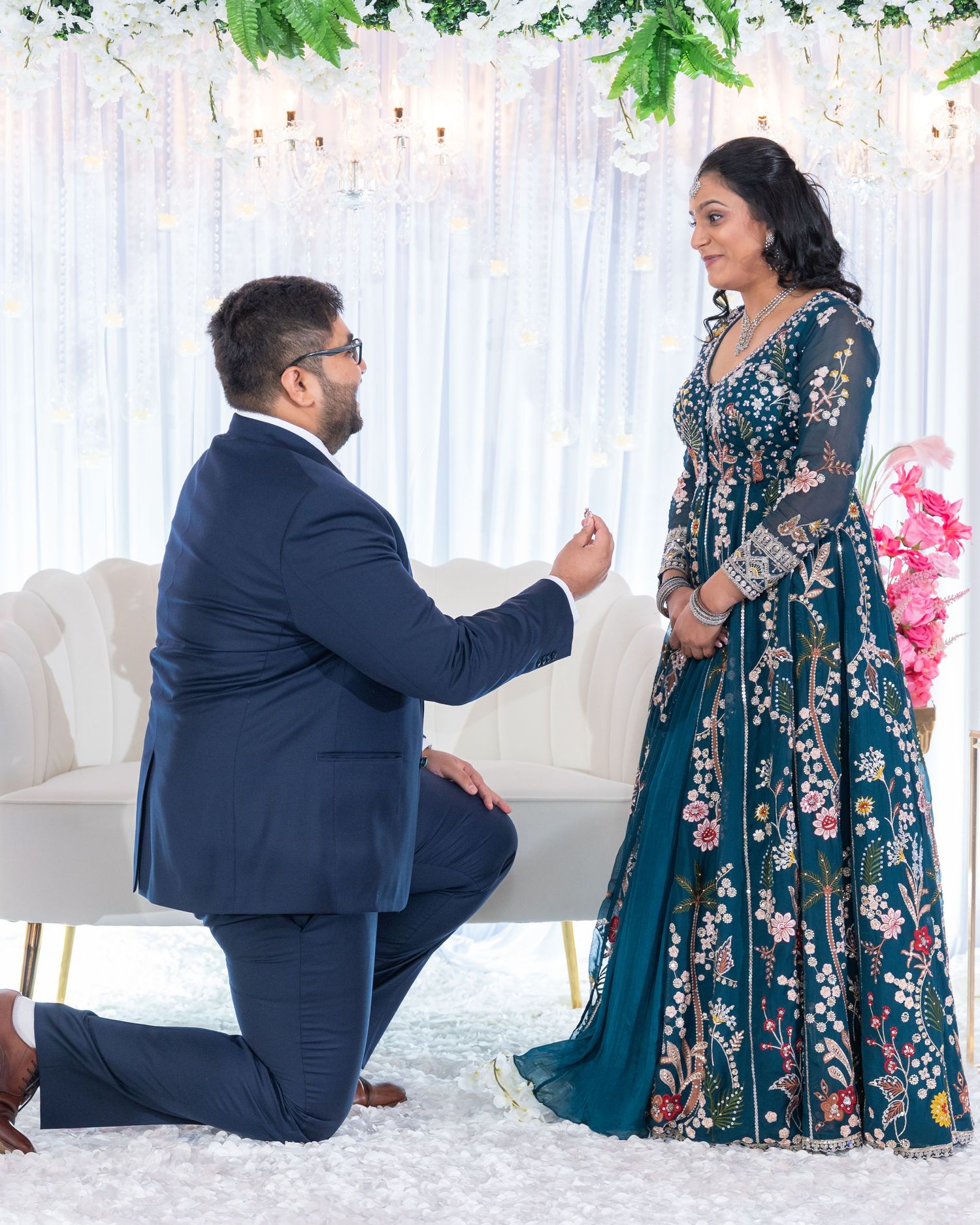 A proposal scene with a floral dress and navy suit against a white backdrop with pink flowers.