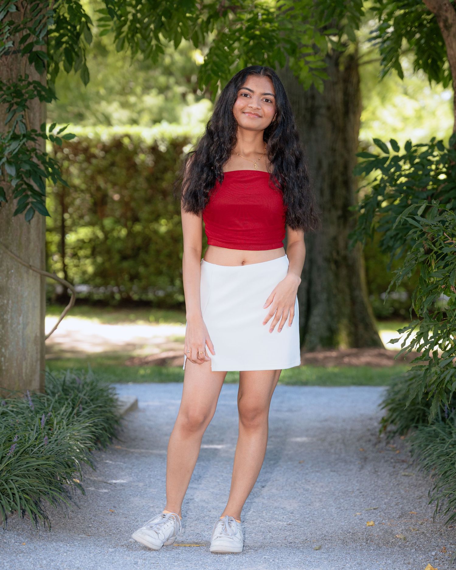 A person in a red top and white skirt poses outdoors on a garden pathway.