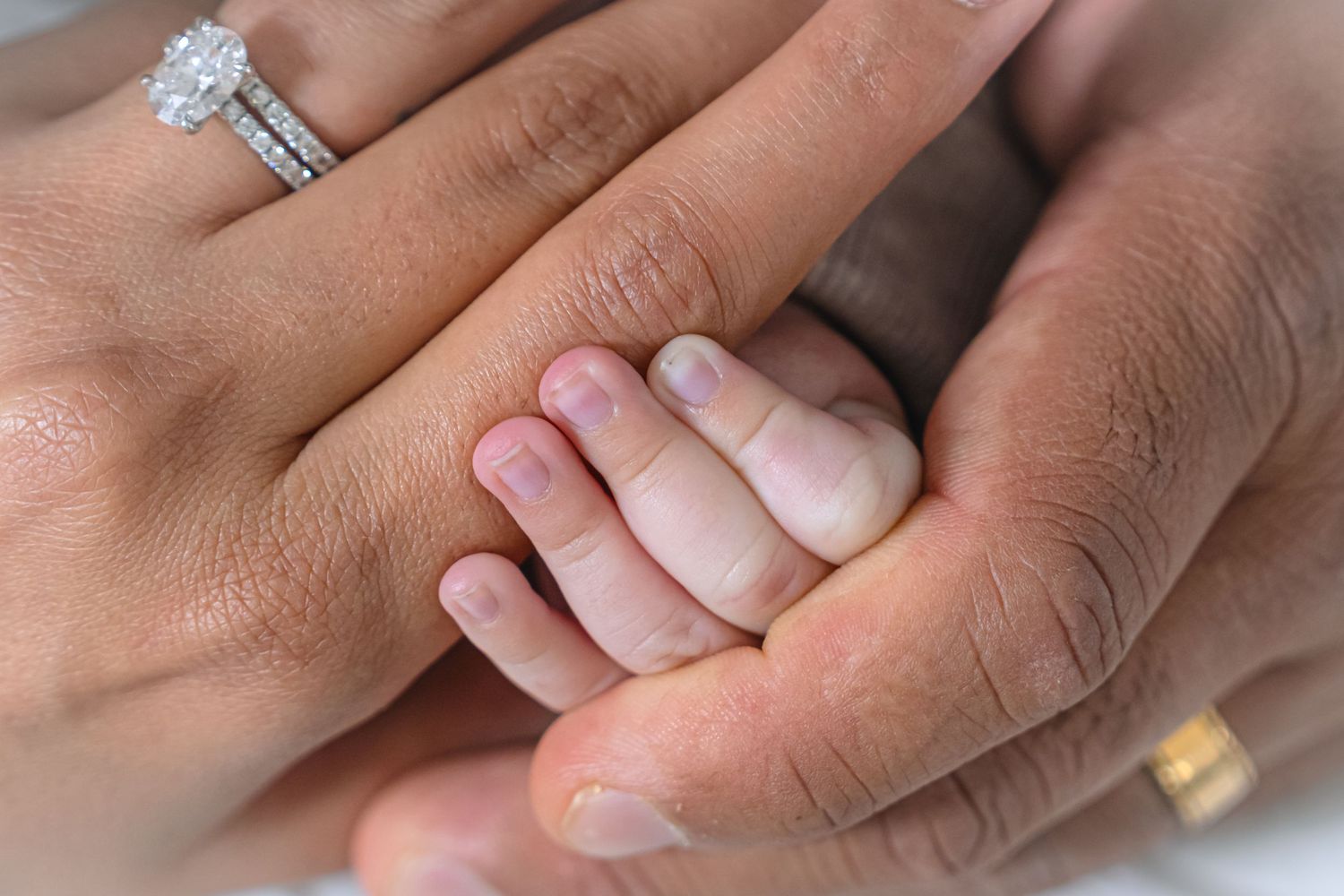 Close-up of newborn baby hand held by parents, mother wearing diamond engagement ring and father with gold wedding band.