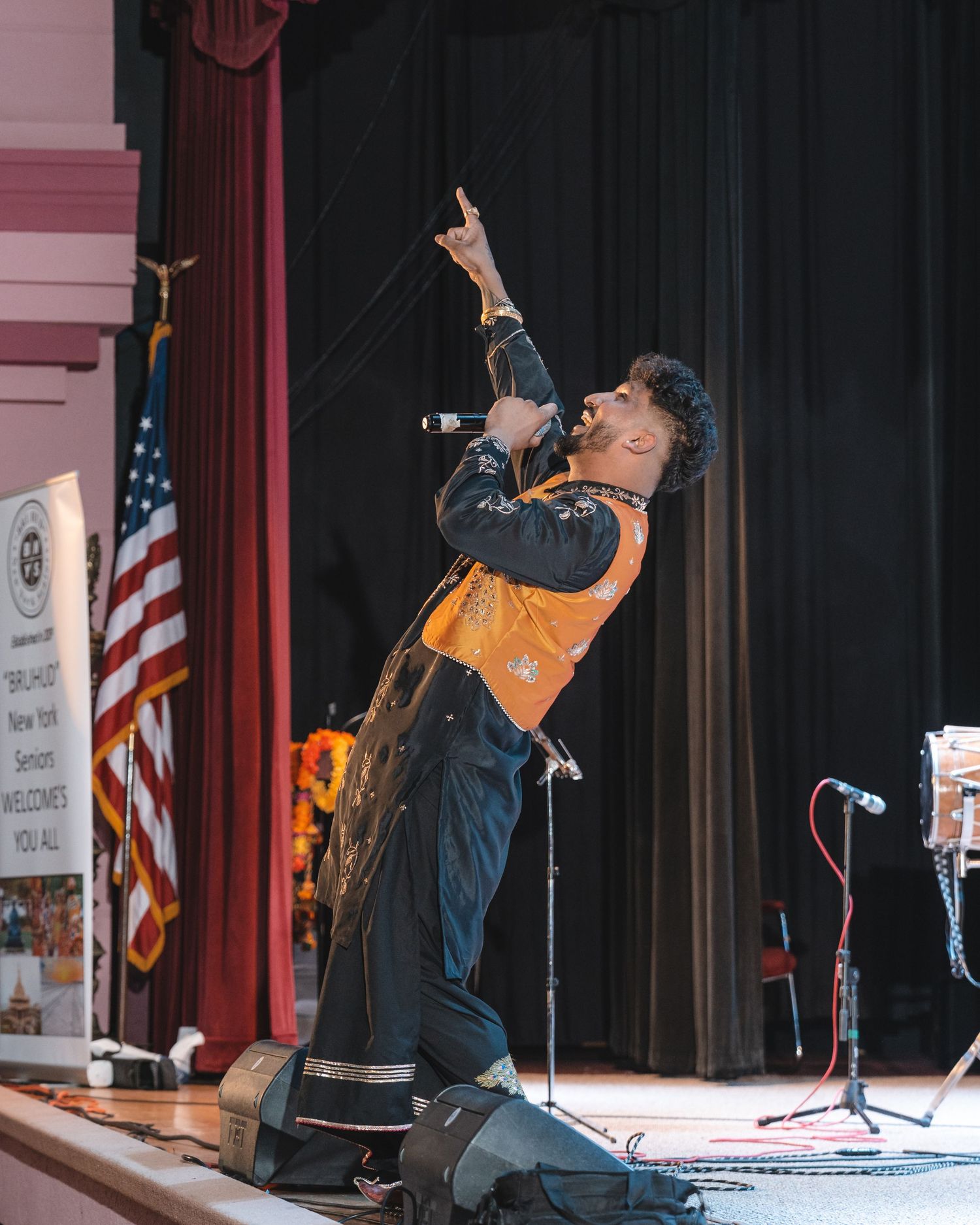 A performer in dark clothing sings passionately on stage with an American flag visible in the background.