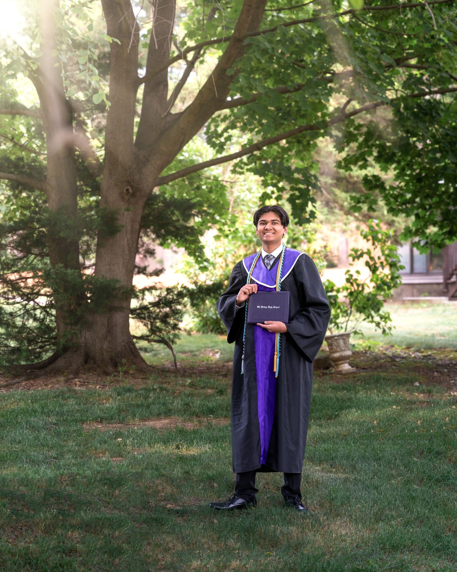 Graduate in academic regalia poses on grassy lawn under large trees.