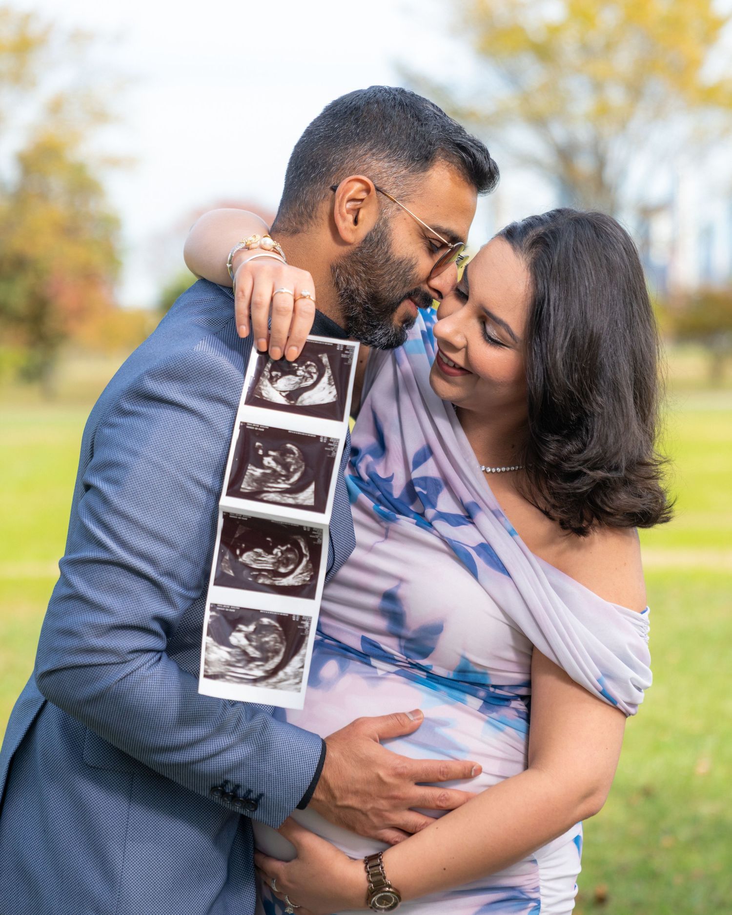 Couple embraces while holding ultrasound photos in a sunny autumn park setting.