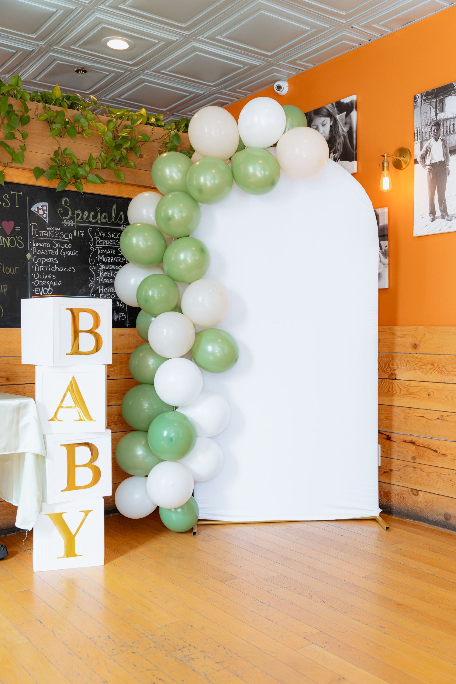 Baby shower party setup with white and sage green balloon arch, decorative letter blocks and hardwood flooring.
