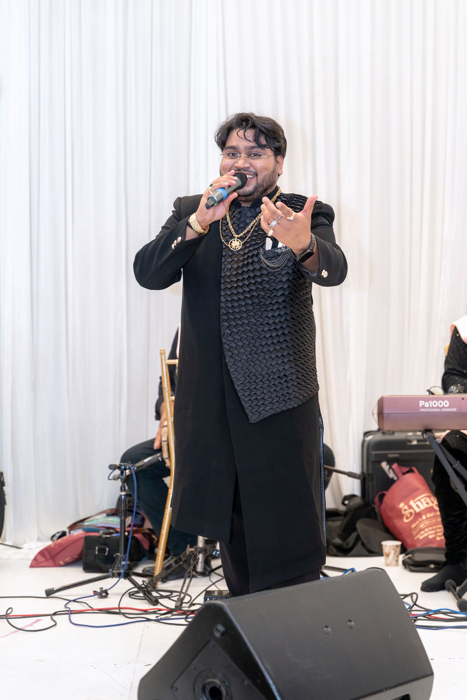 A performer in black attire sings passionately on stage with sound equipment and white curtains in the background.