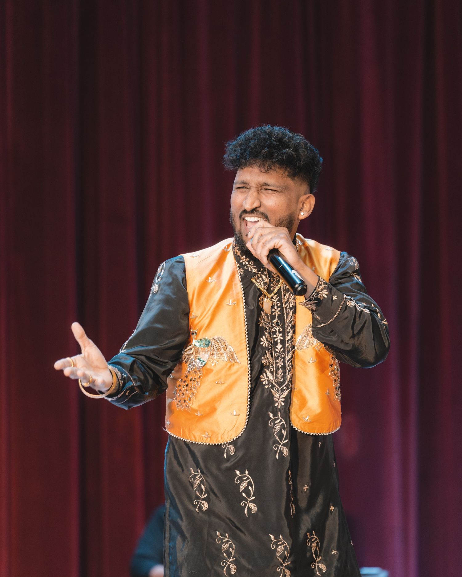 Performer in black and orange vest performs energetic dance moves on stage with red curtain backdrop.